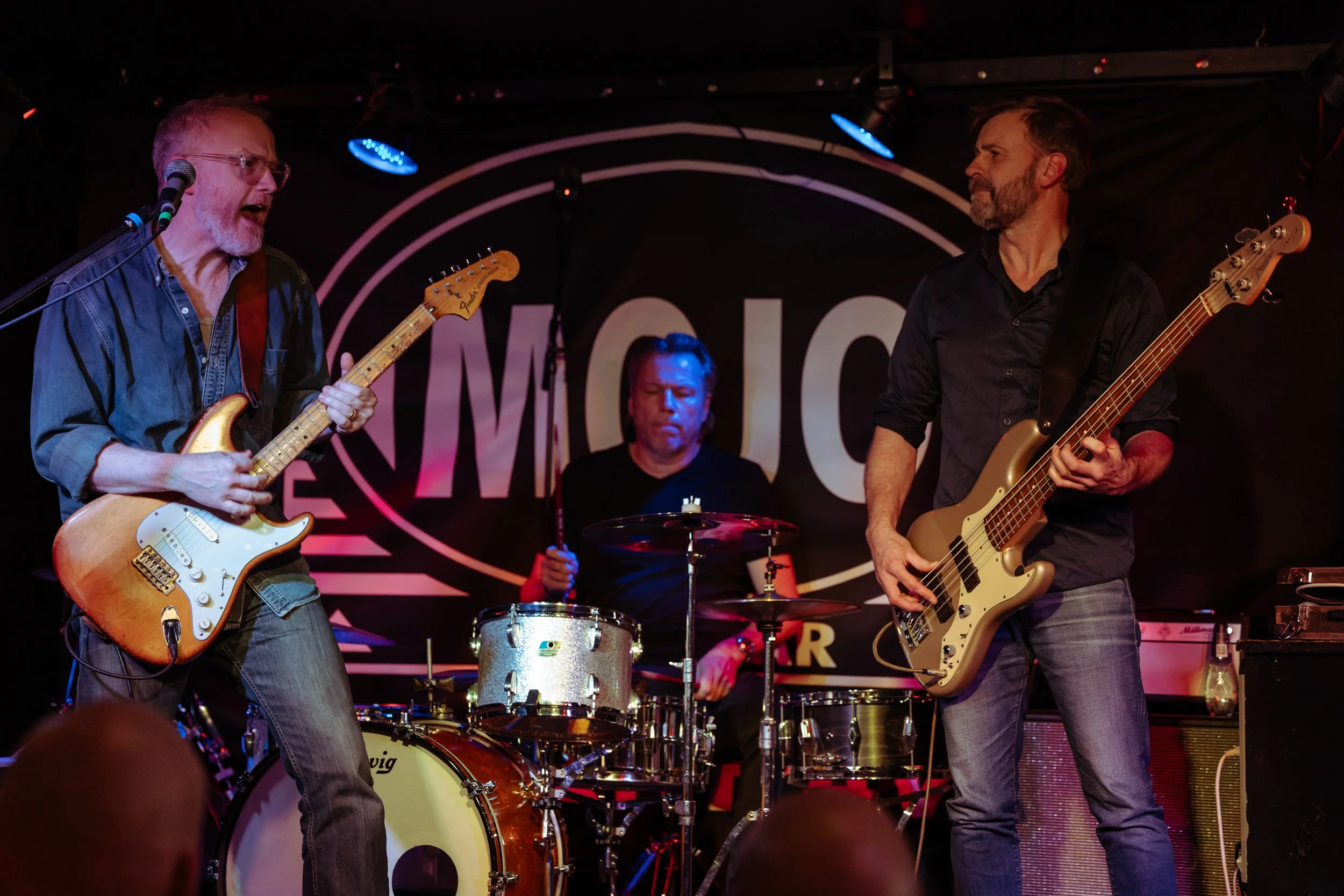 A band performing on stage with two guitarists in the foreground and a drummer in the background. The guitarist on the left is an older man with glasses, a beard, and short hair, playing a Fender Stratocaster. The guitarist on the right is a middle-aged man with a beard, playing a bass guitar. The drummer is a middle-aged man with dark hair, playing drums. The band is on a stage with a dark background and stage lighting.
