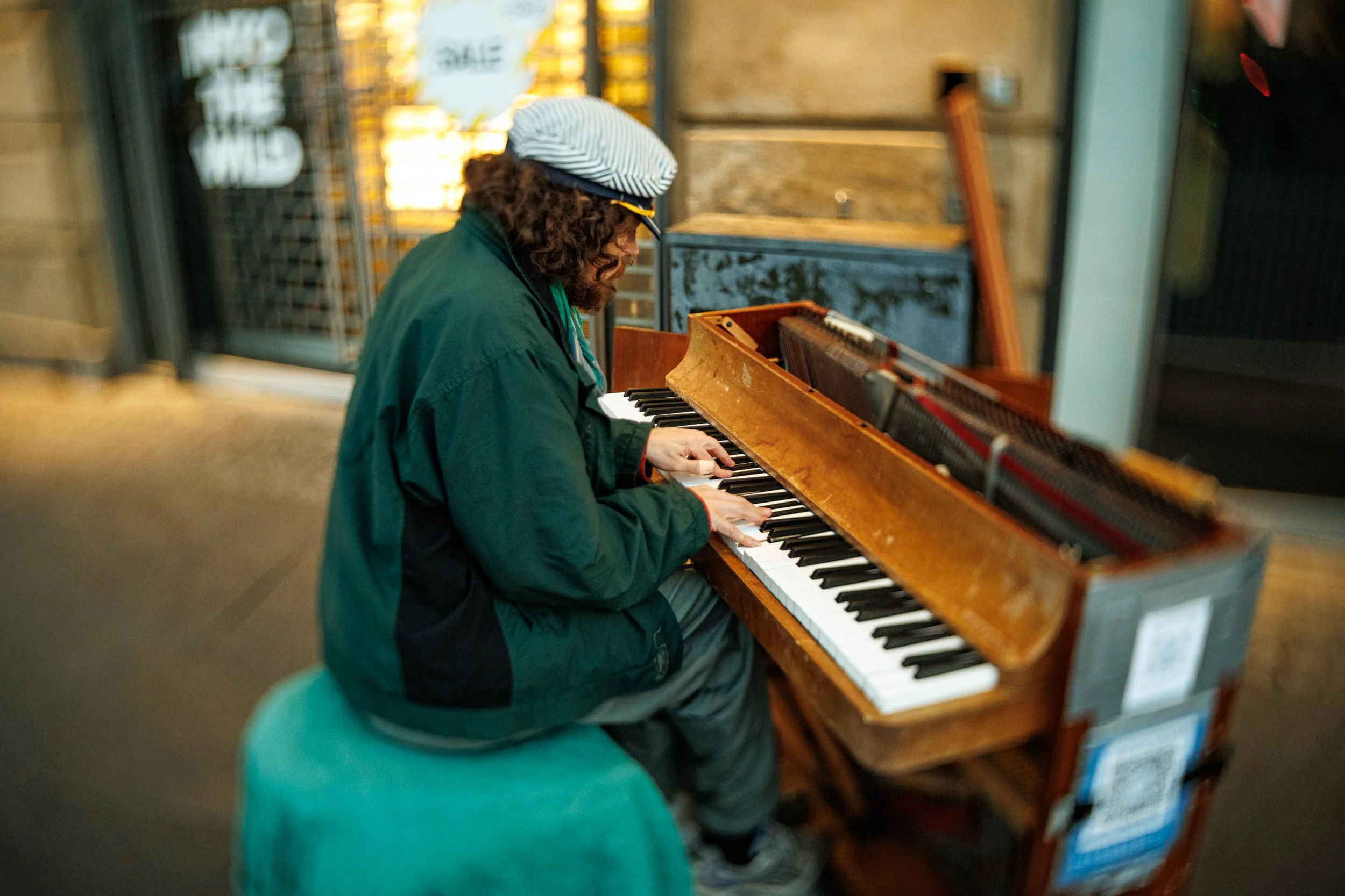 A person with curly hair, wearing a striped hat and a green jacket, playing an old upright piano inside a building.