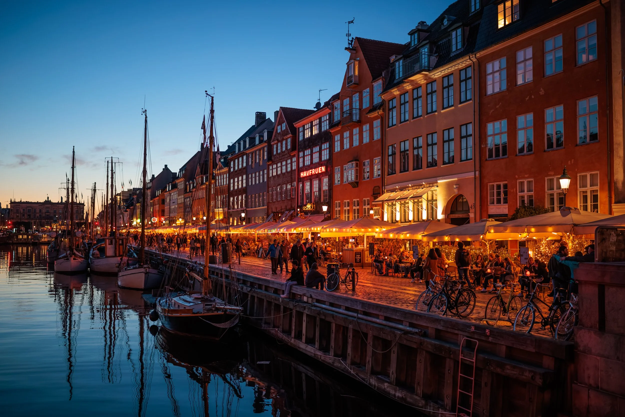 Colorful waterfront street scene at dusk with boats docked in the harbor, buildings with illuminated windows, and people sitting at outdoor cafes.