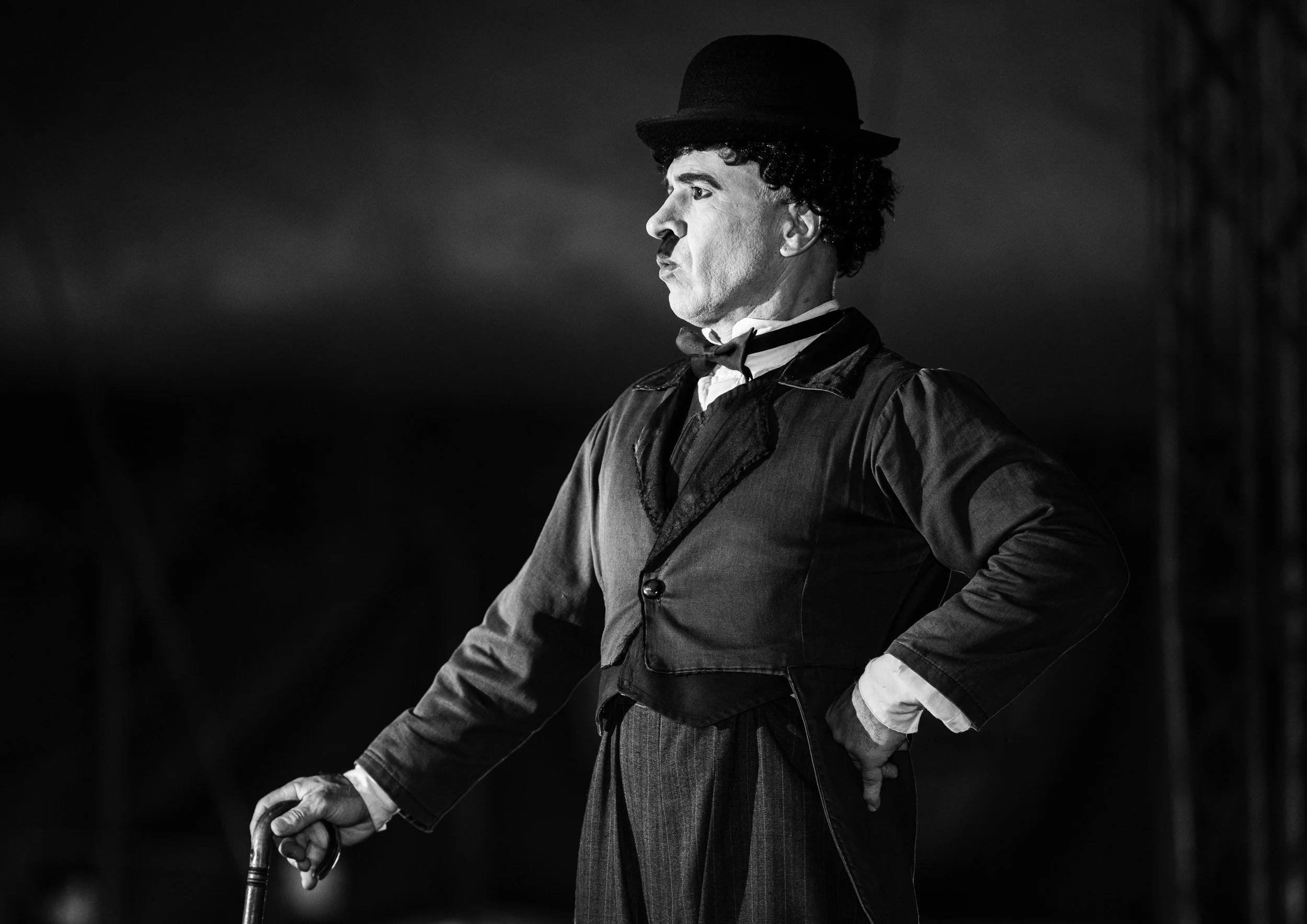 Black and white photo of Charlie Chaplin dressed as the Tramp, standing sideways with arms akimbo, wearing a bowler hat, a suit, and holding a cane.