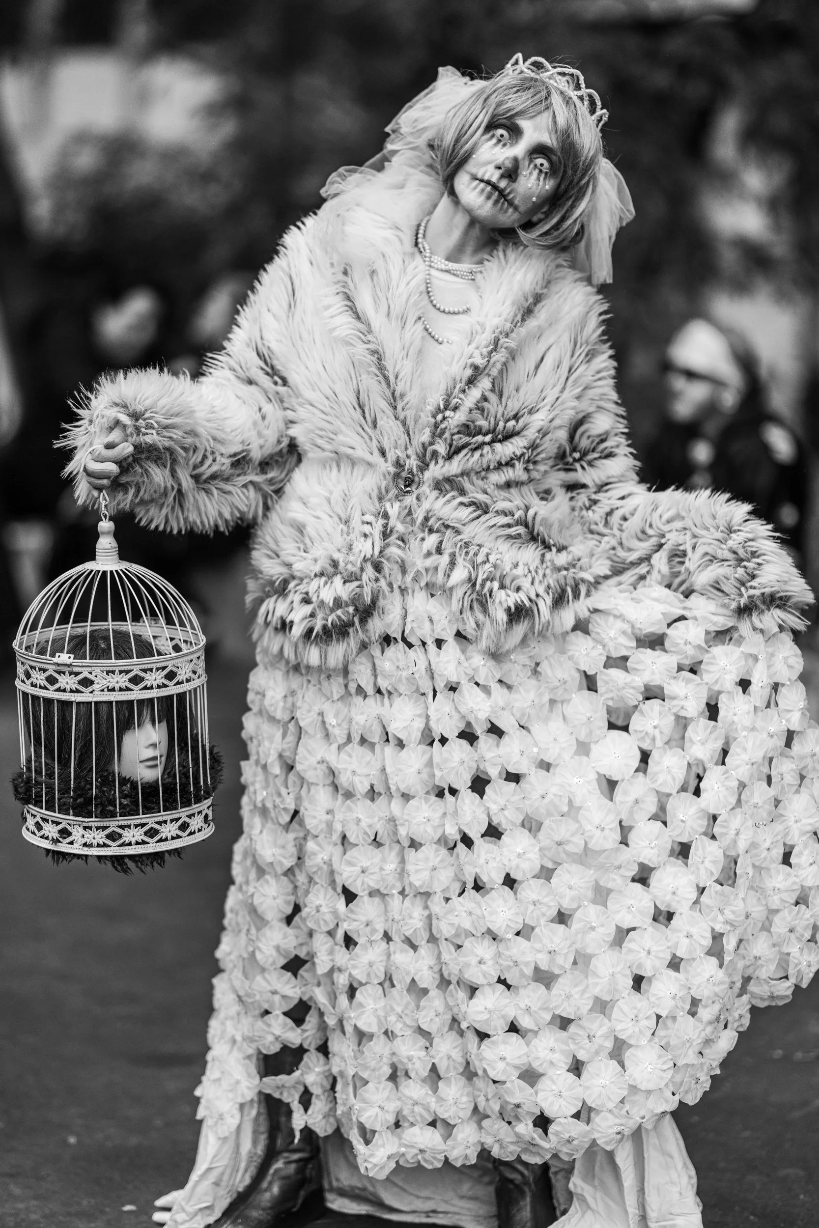 A person dressed in a costume resembling a doll or mannequin, wearing a fur coat and a dress made of flowers. They have makeup to look like a skeletal face with tears, and are surrounded by a crowd, with one person in the background wearing glasses a