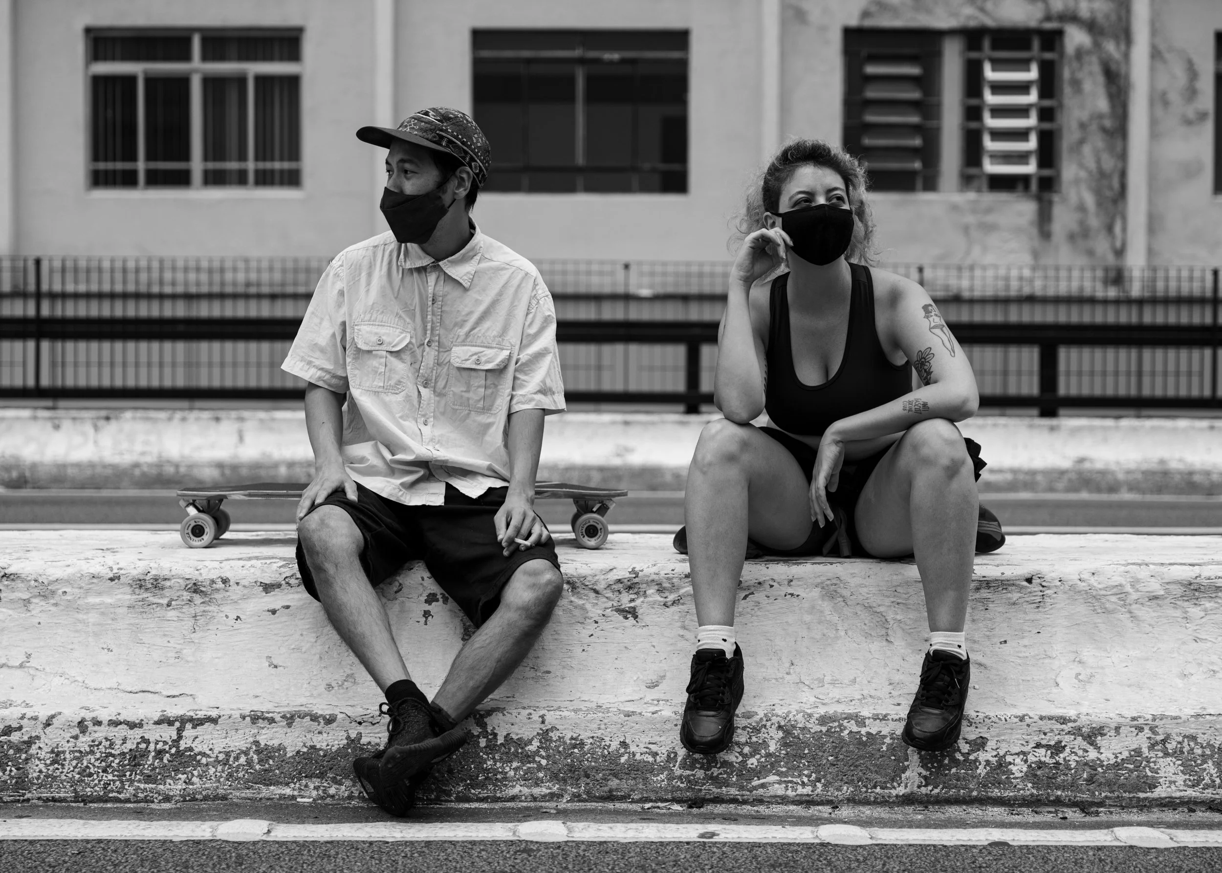 Two people sitting on a ledge outdoors, wearing face masks. The person on the left is a man with a skateboard, wearing a cap and casual clothing. The person on the right is a woman with curly hair, Tattoos on her arms, wearing a tank top and shorts. 