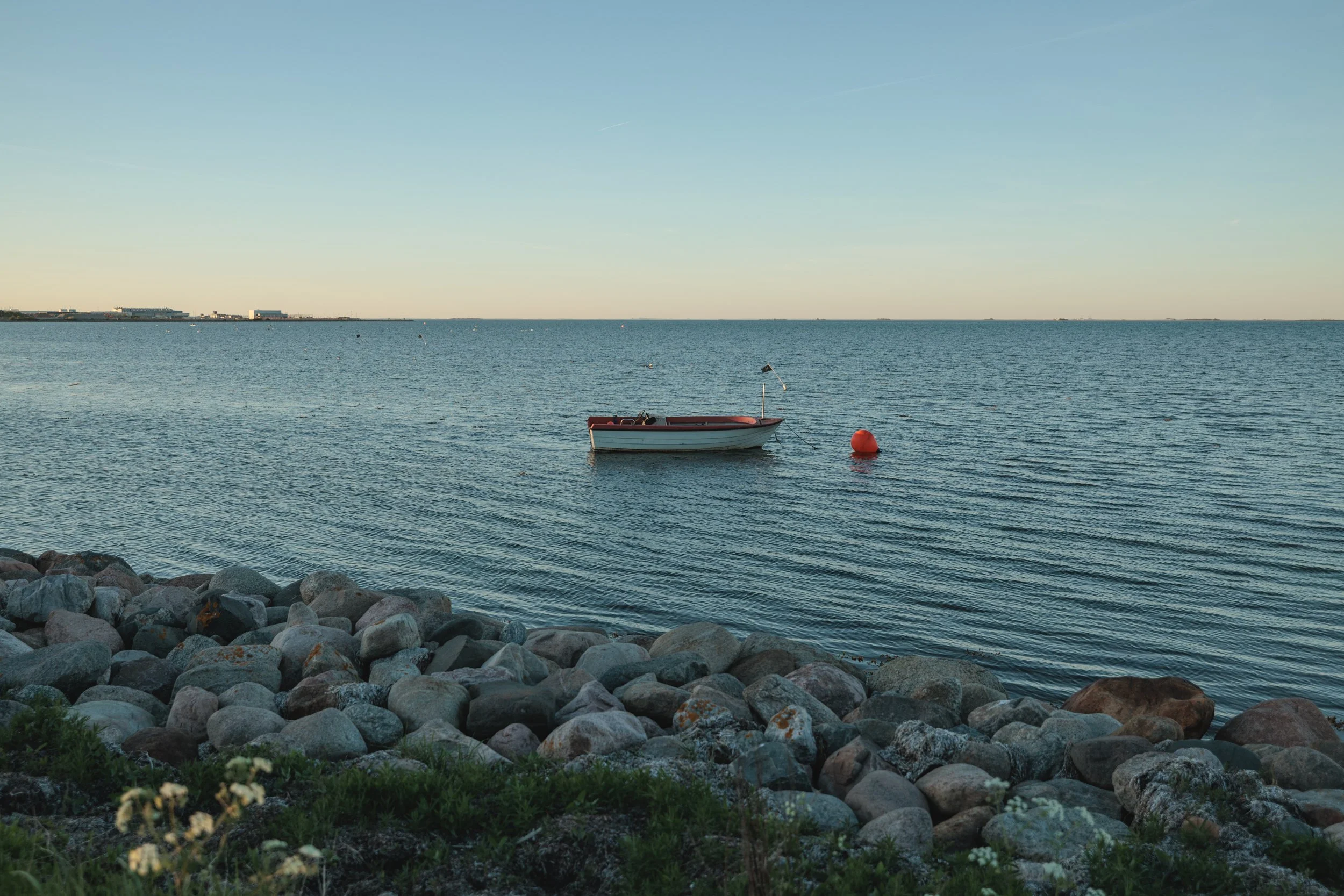 A small boat floating on calm water near a rocky shoreline, with a clear sky and industrial buildings in the distance.