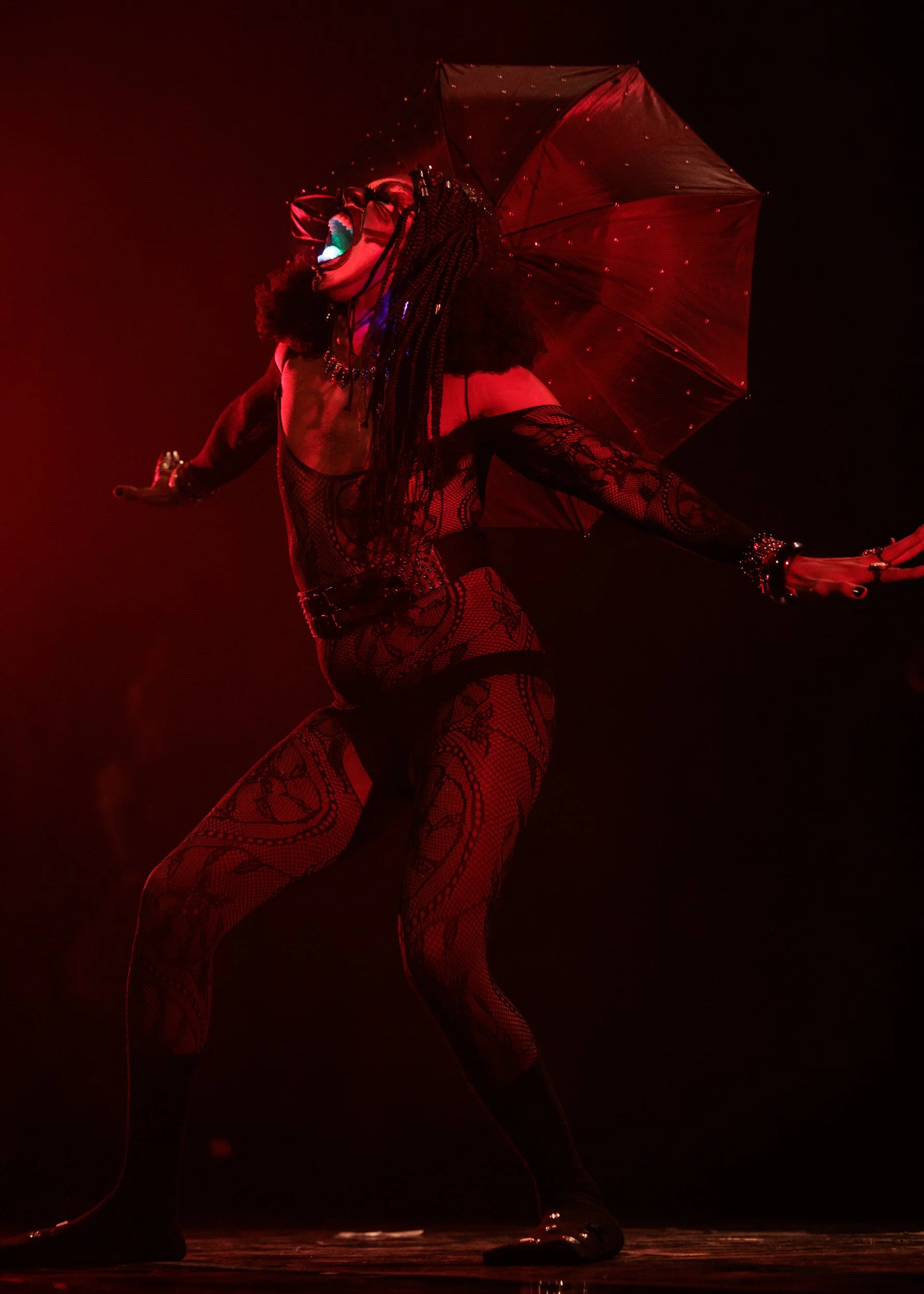 A performer on stage wearing fishnet tights, multiple bracelets, and a necklace, holding a transparent umbrella with small lights, illuminated by a red stage light.