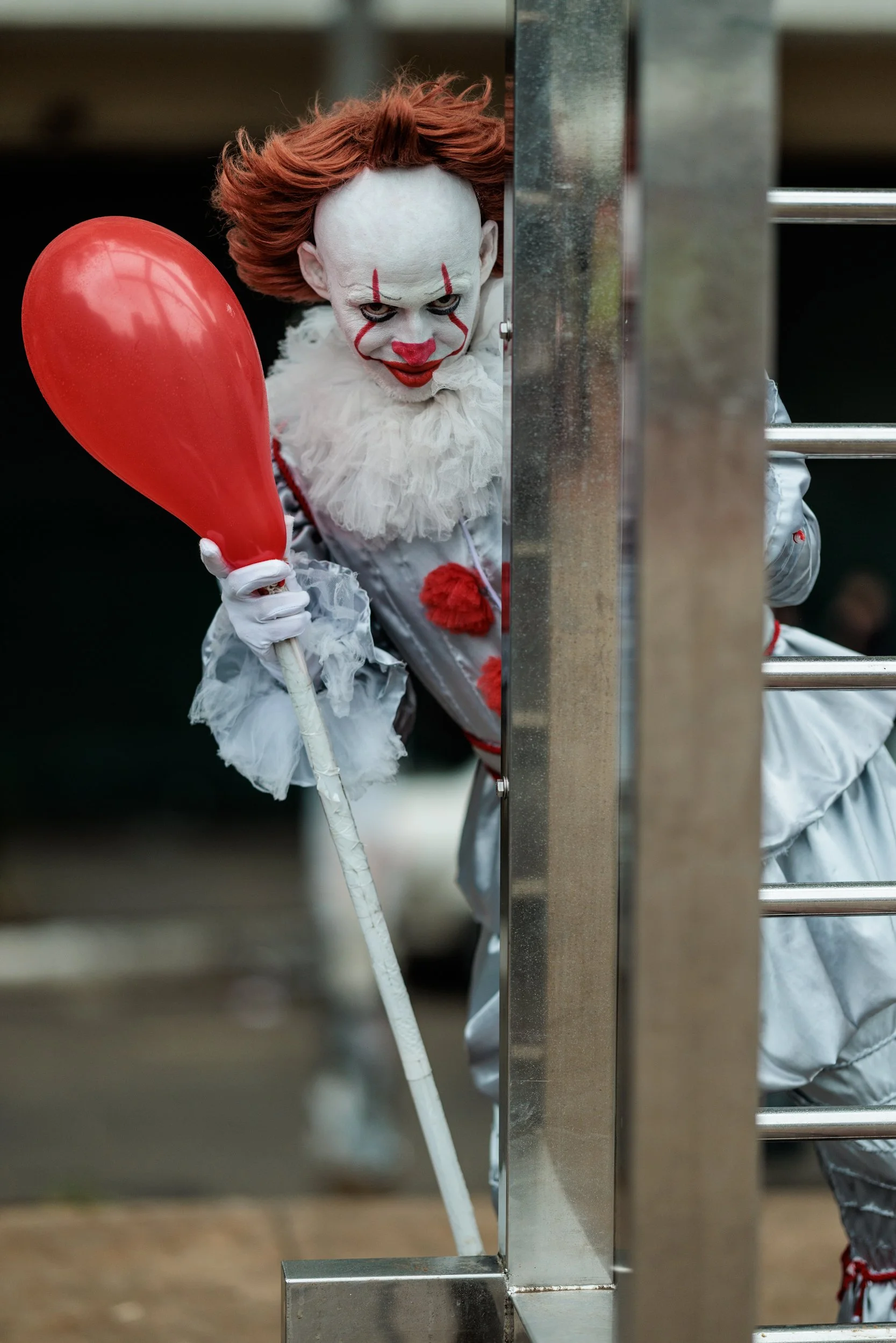 Person dressed as a scary clown with white face paint, red makeup, and orange hair, holding a red balloon and peeking through a metal barrier.