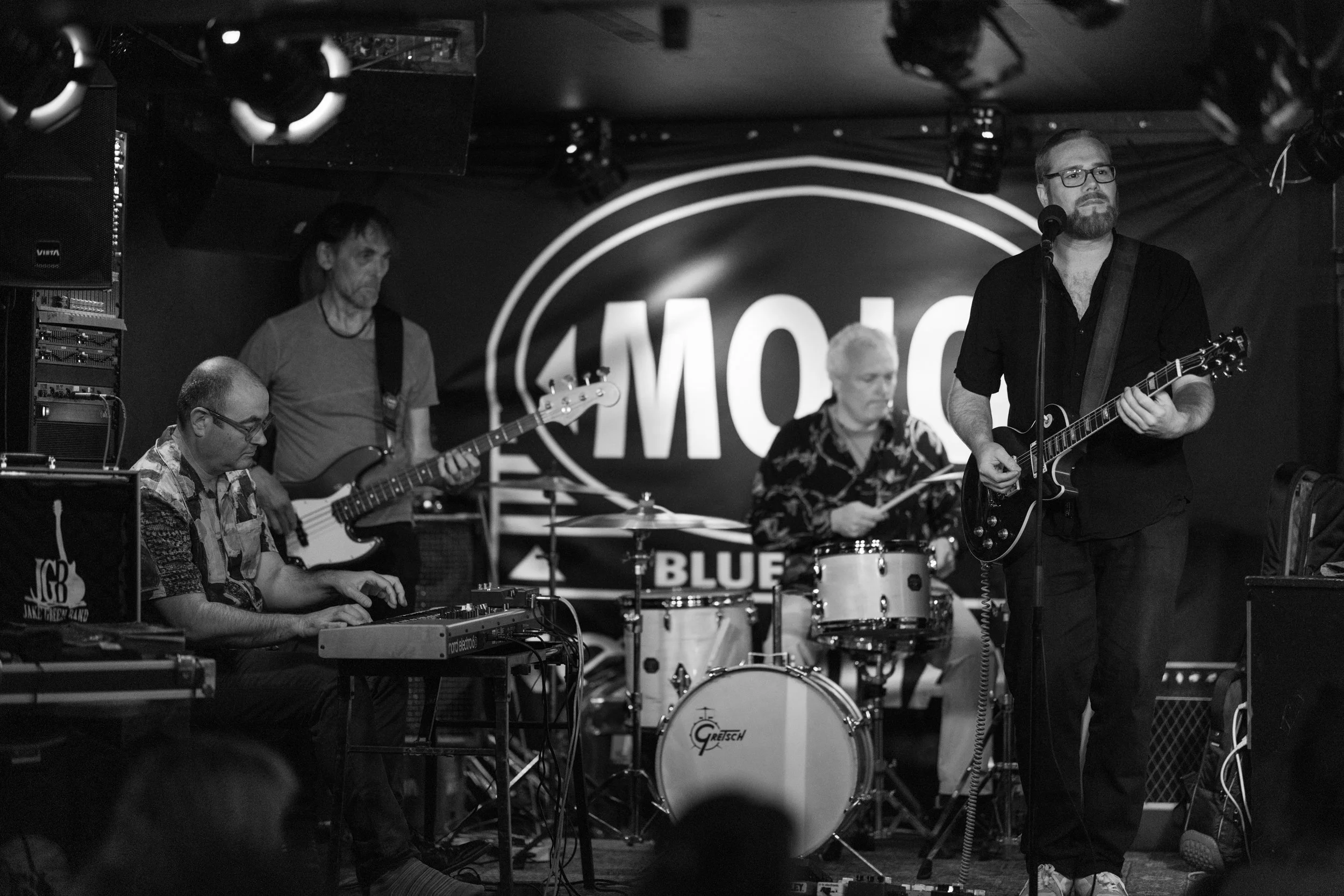 A black-and-white photo of a live band performing on stage. There are five musicians with various instruments, including a keyboard, bass guitar, drum set, and an electric guitar. The backdrop features a circular logo with the word 'MOTION' visible. 