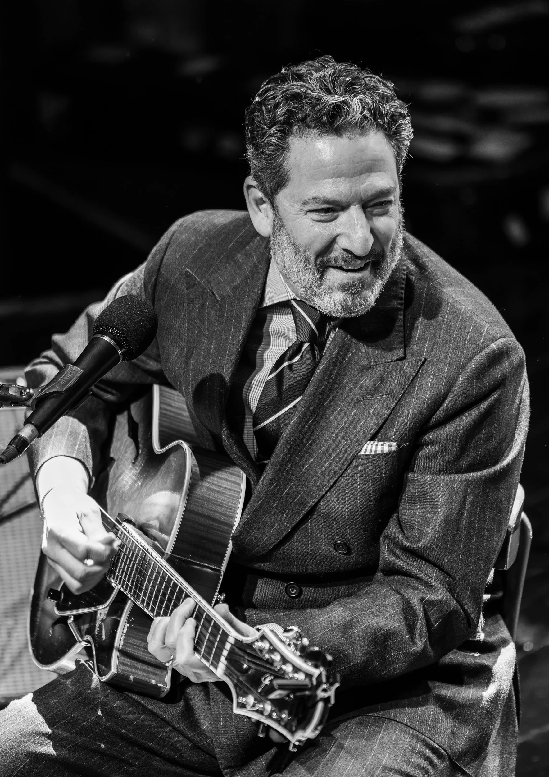 Black and white photo of a man with curly hair and a beard playing an acoustic guitar and singing into a microphone, dressed in a pinstripe suit.
