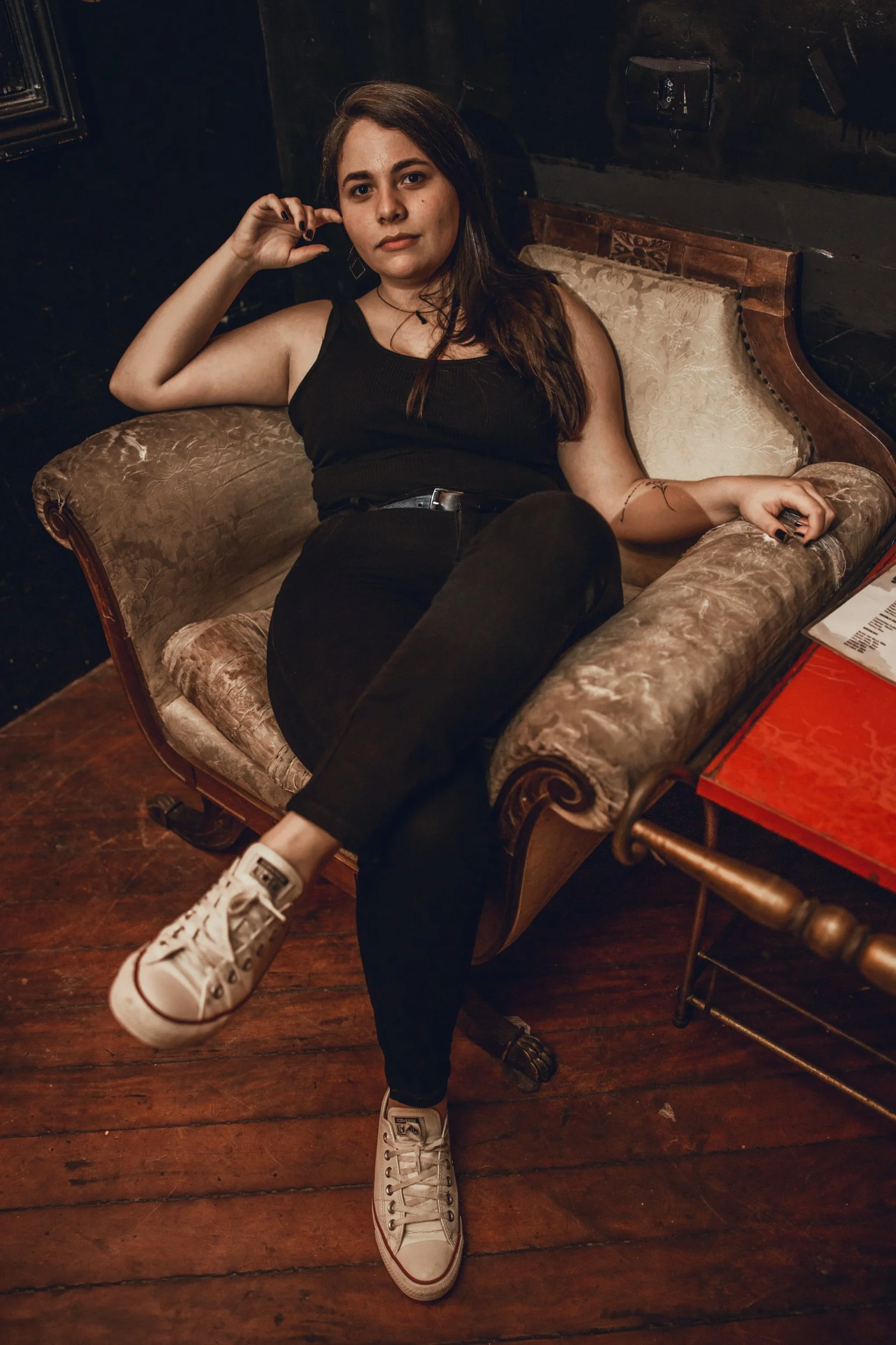 A young woman with long dark hair and light skin is sitting on a vintage sofa with floral upholstery in a dimly lit room. She wears a black sleeveless top, black pants, and white sneakers. She has a relaxed expression and is resting her head on her l