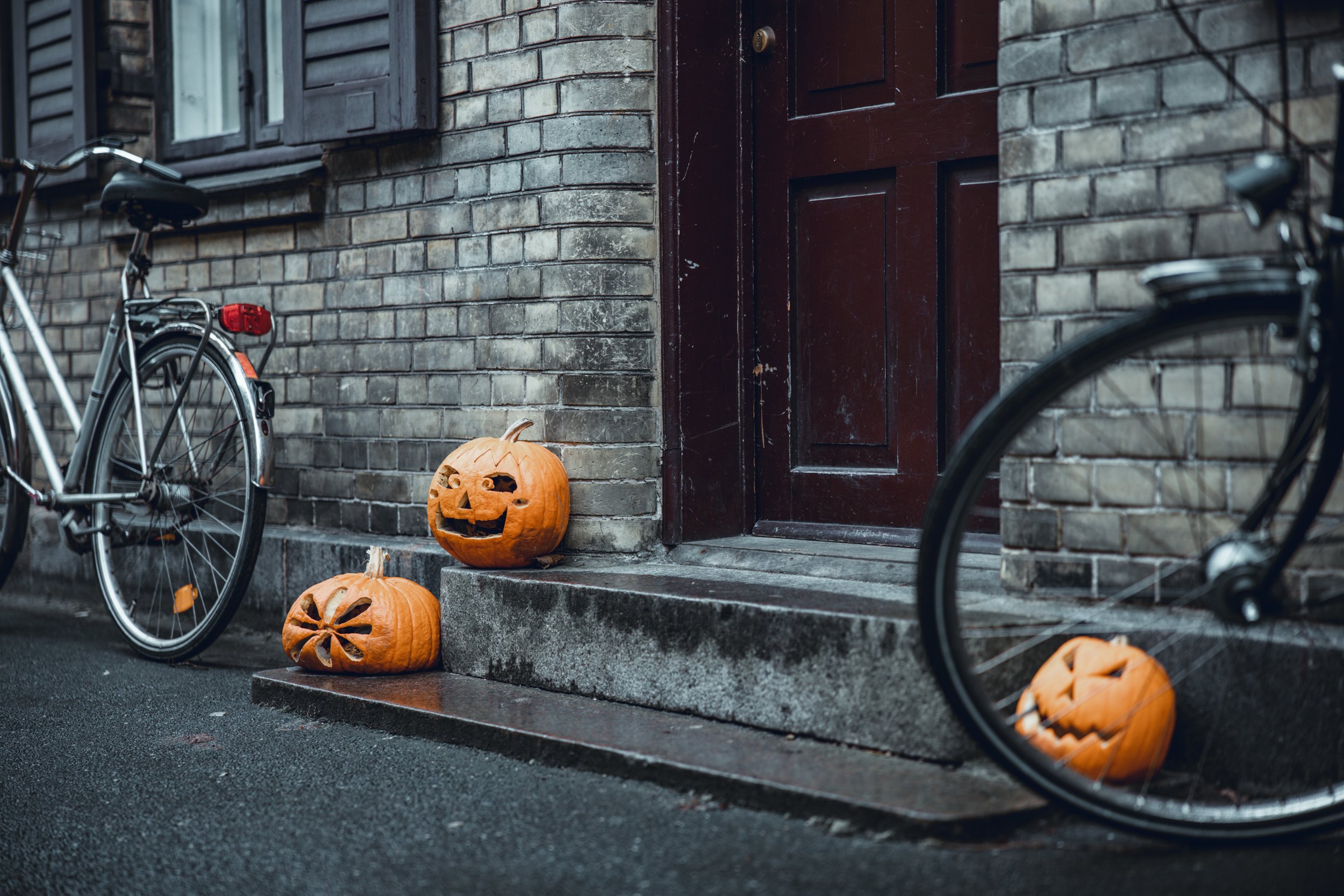 Halloween pumpkins with carved faces placed on the doorstep of a brick building, with two bicycles parked nearby.