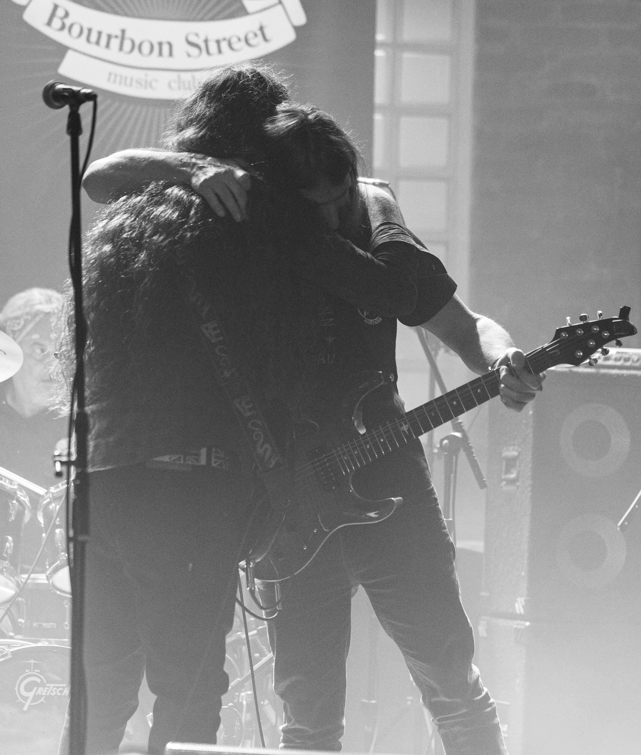 Black and white photo of two people hugging on stage with musical equipment, a microphone, and a sign for Bourbon Street music club in the background.