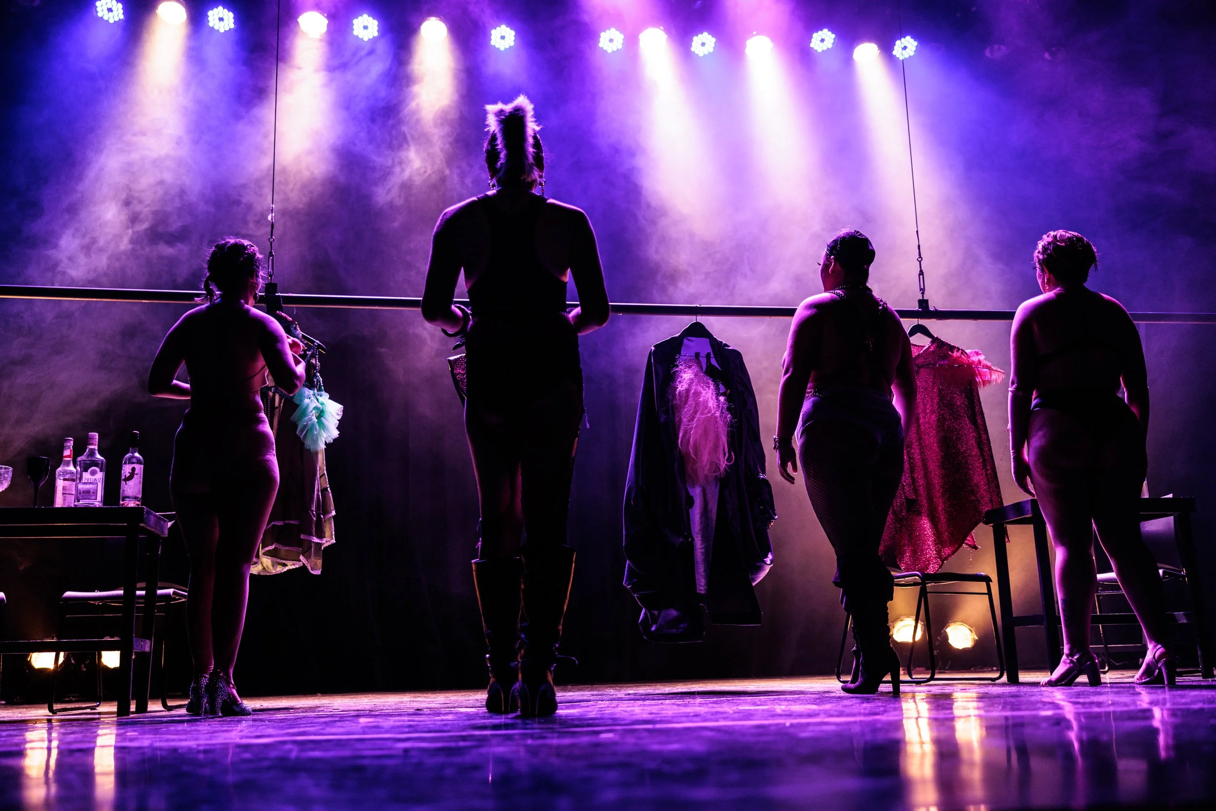 Silhouettes of four women standing on a stage, backlit by purple and blue stage lights, with costumes and clothing hanging on a rack, and bottles on a table in front.