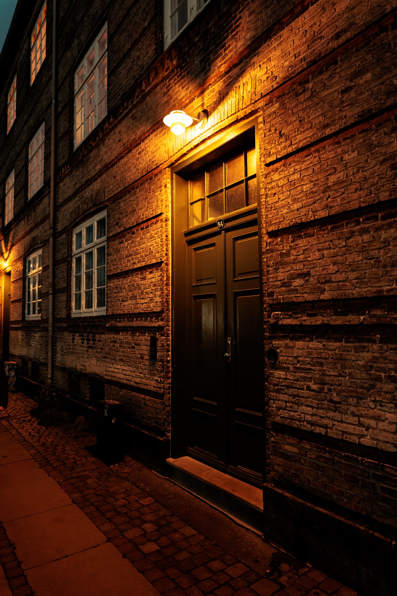 Nighttime view of a brick building with a black door, illuminated by an exterior wall-mounted light. Windows are visible on the upper floors, and cobblestone pavement runs along the sidewalk.