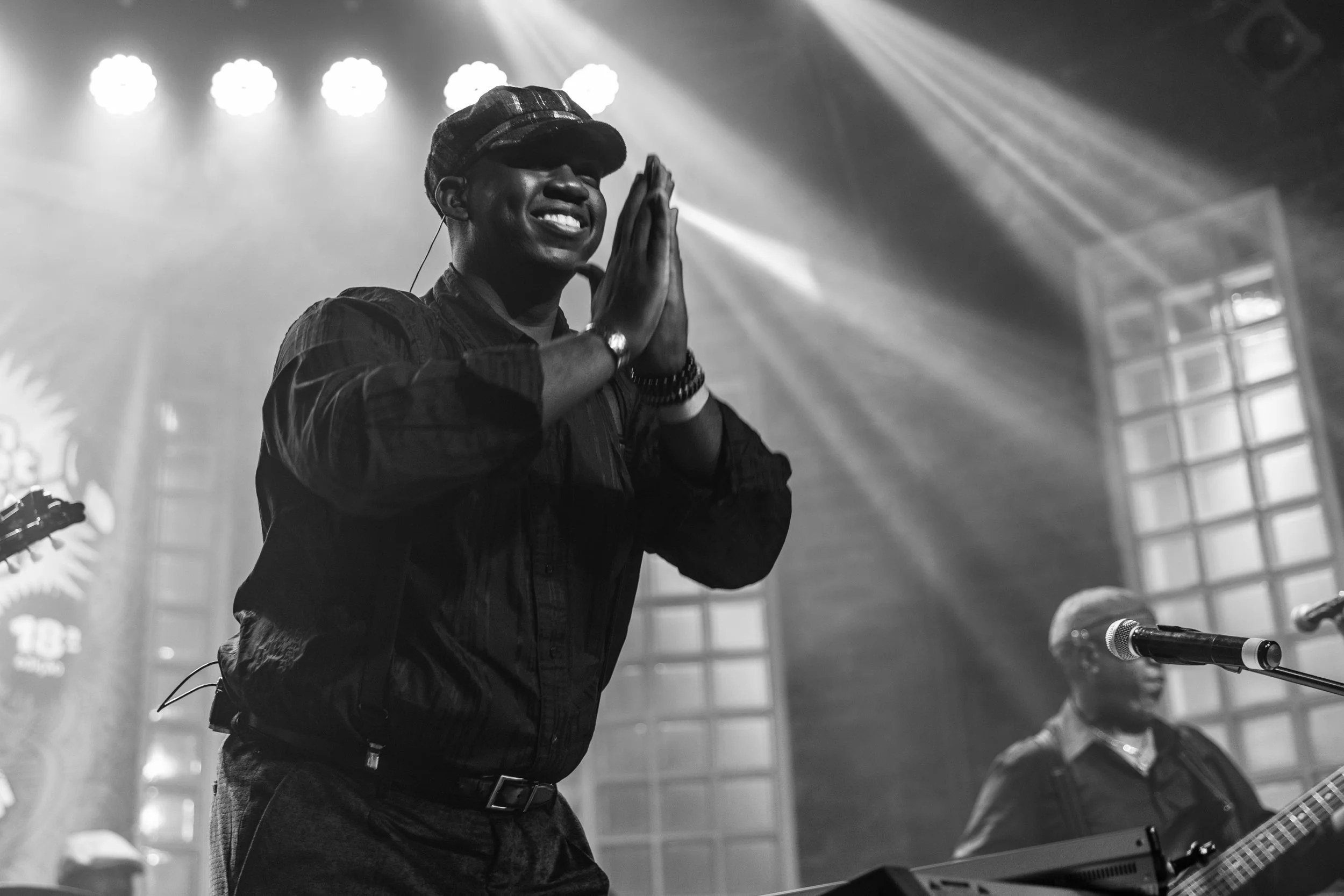 A person wearing a cap and jacket, with hands pressed together in a prayer or greeting gesture, smiling while performing on stage with bright lights shining behind them.