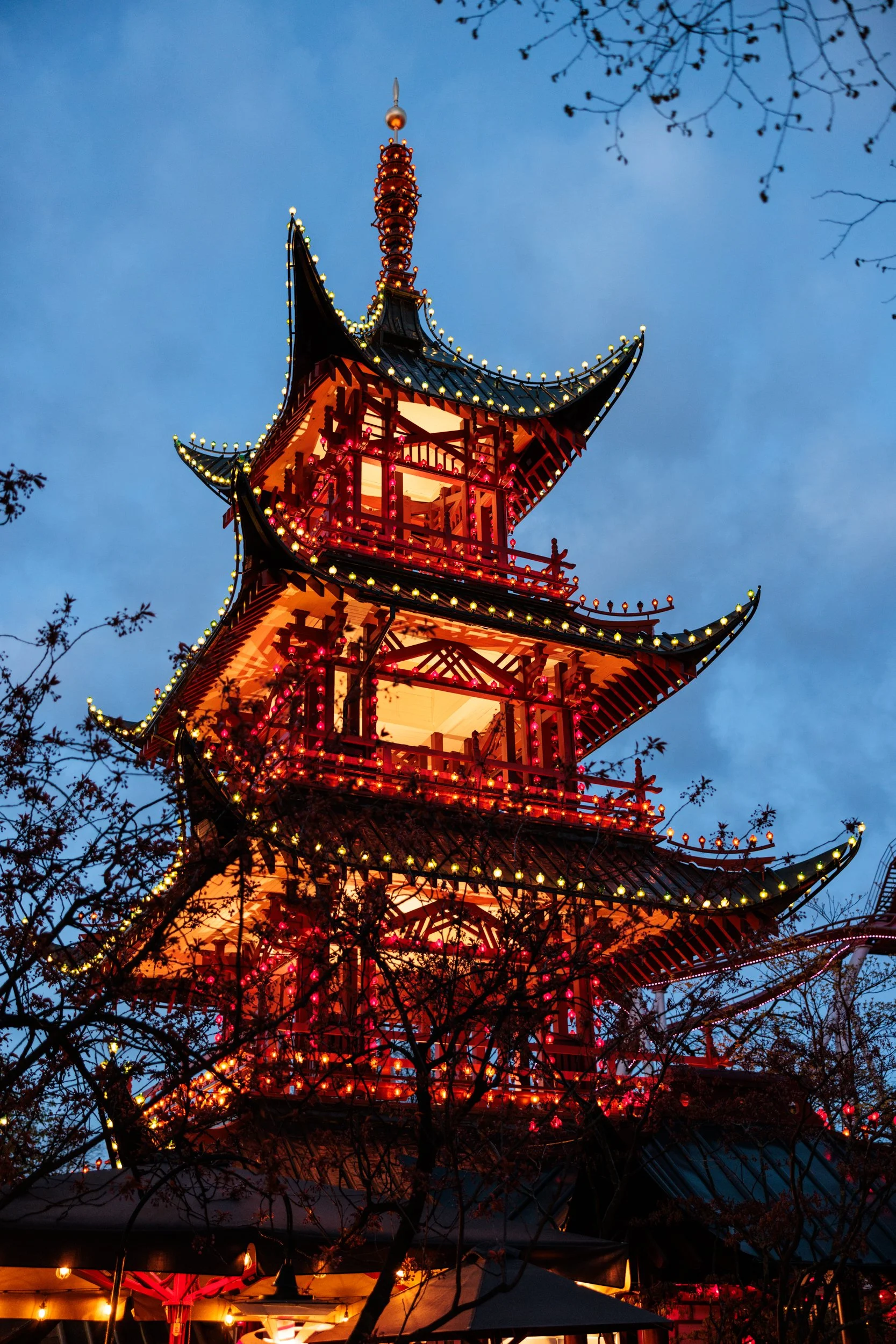 A multi-tiered traditional Asian pagoda with ornate roof details, adorned with multicolored lights against a dusk sky, surrounded by tree branches.