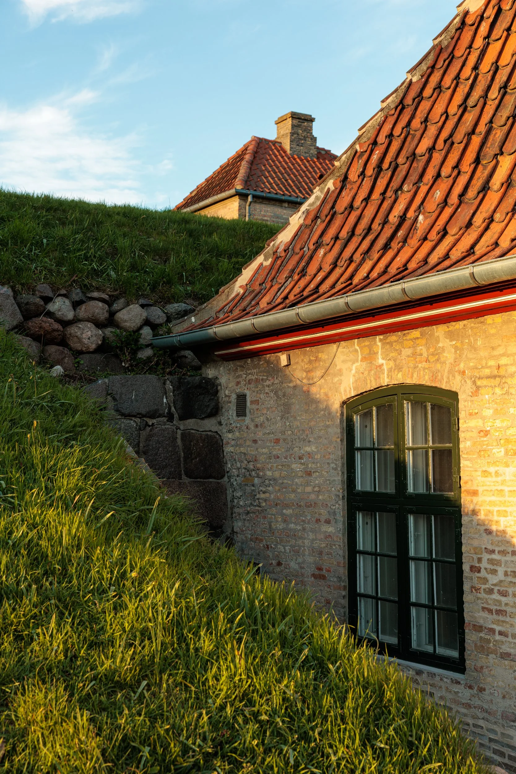 A rustic brick house with a tiled roof and large window, situated next to a grassy hill with rocks, under a blue sky with clouds.
