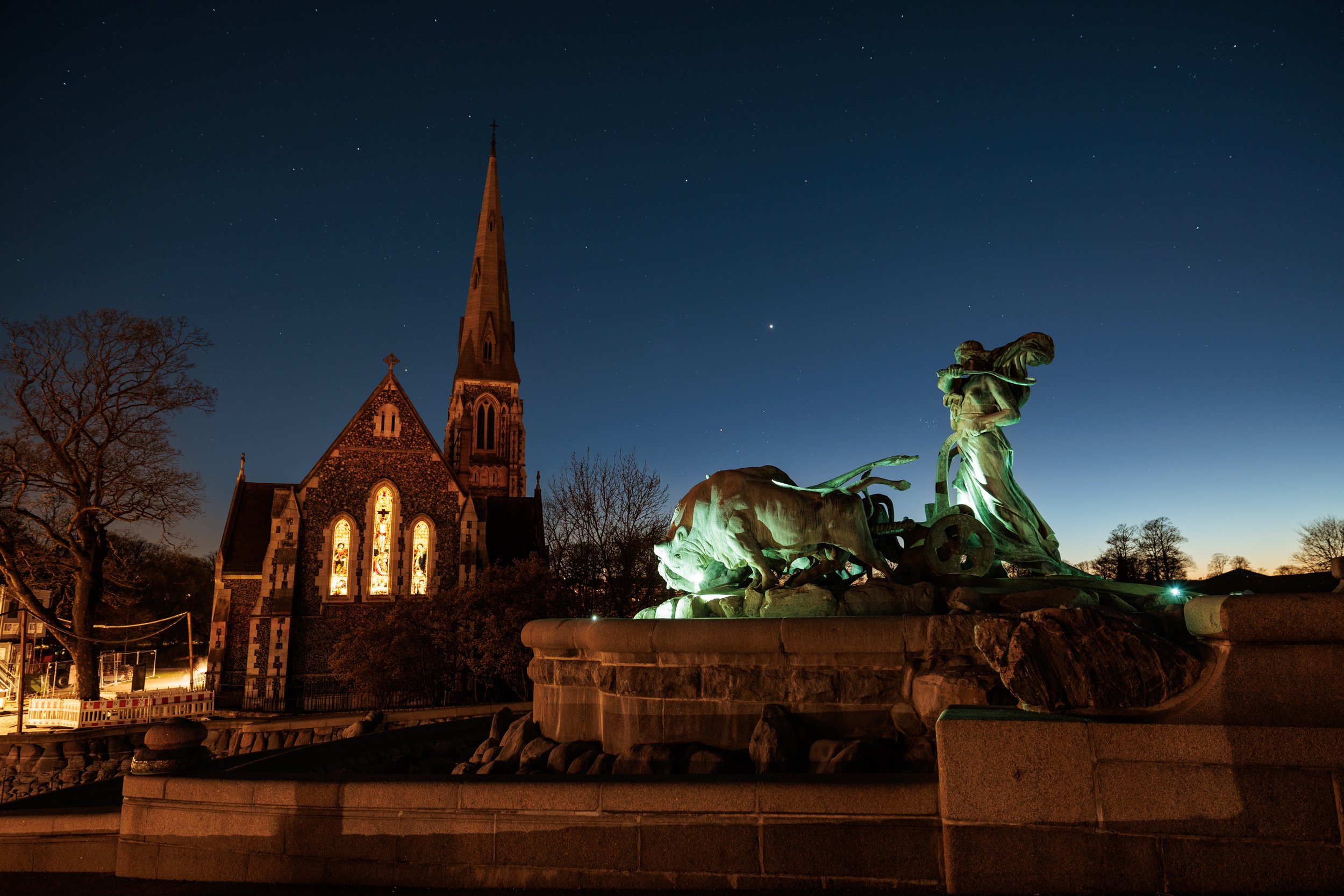 Nighttime scene of a church with stained glass windows and a tall steeple. In front of the church, there is a monument featuring a chariot drawn by two horses and driven by a figure, all illuminated with green lights. The sky above is dark with visib