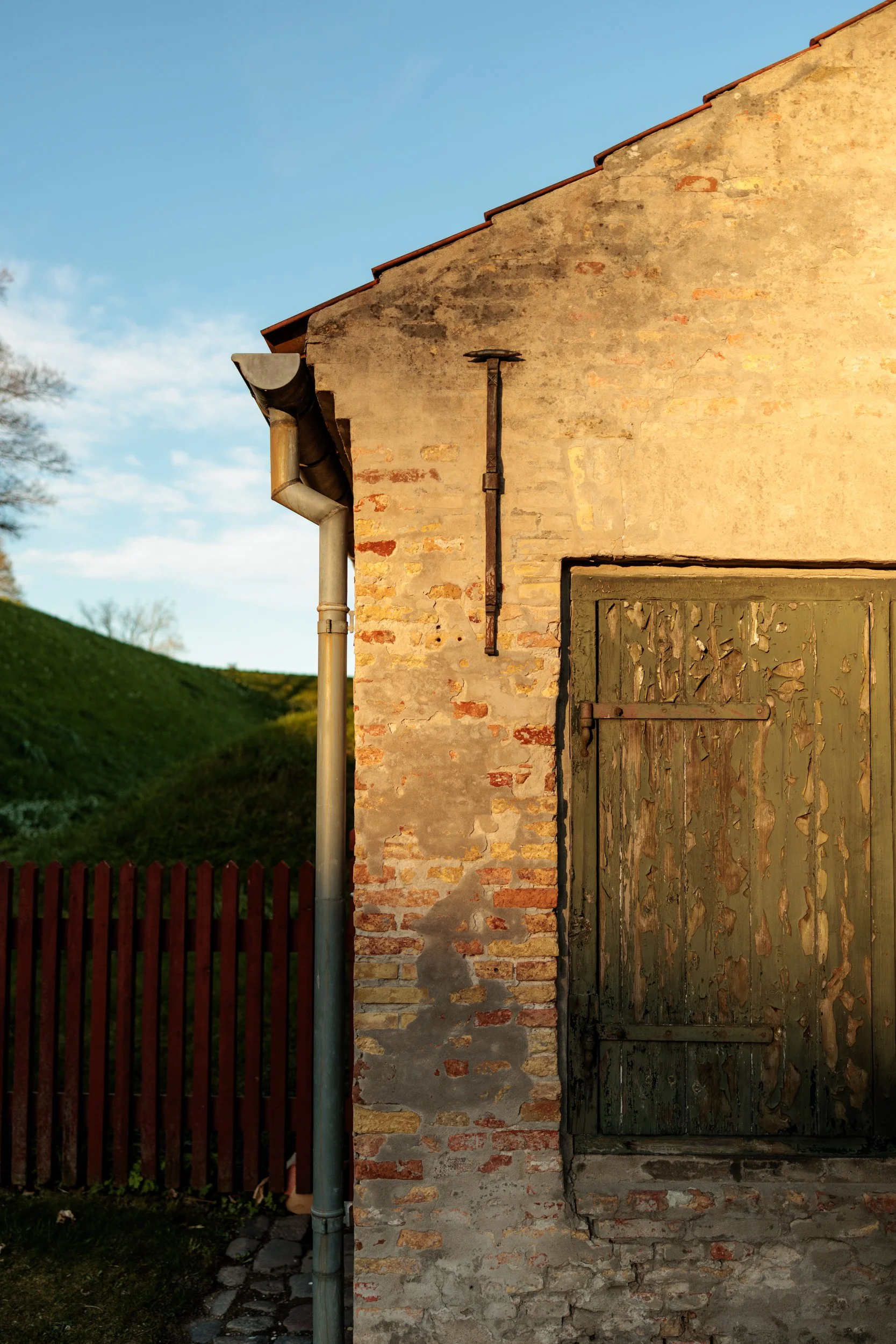Old brick building with weathered green wooden door, rusted iron handle, and old metal pipes on the side, surrounded by a red picket fence and set against a clear sky with green hills in the background.