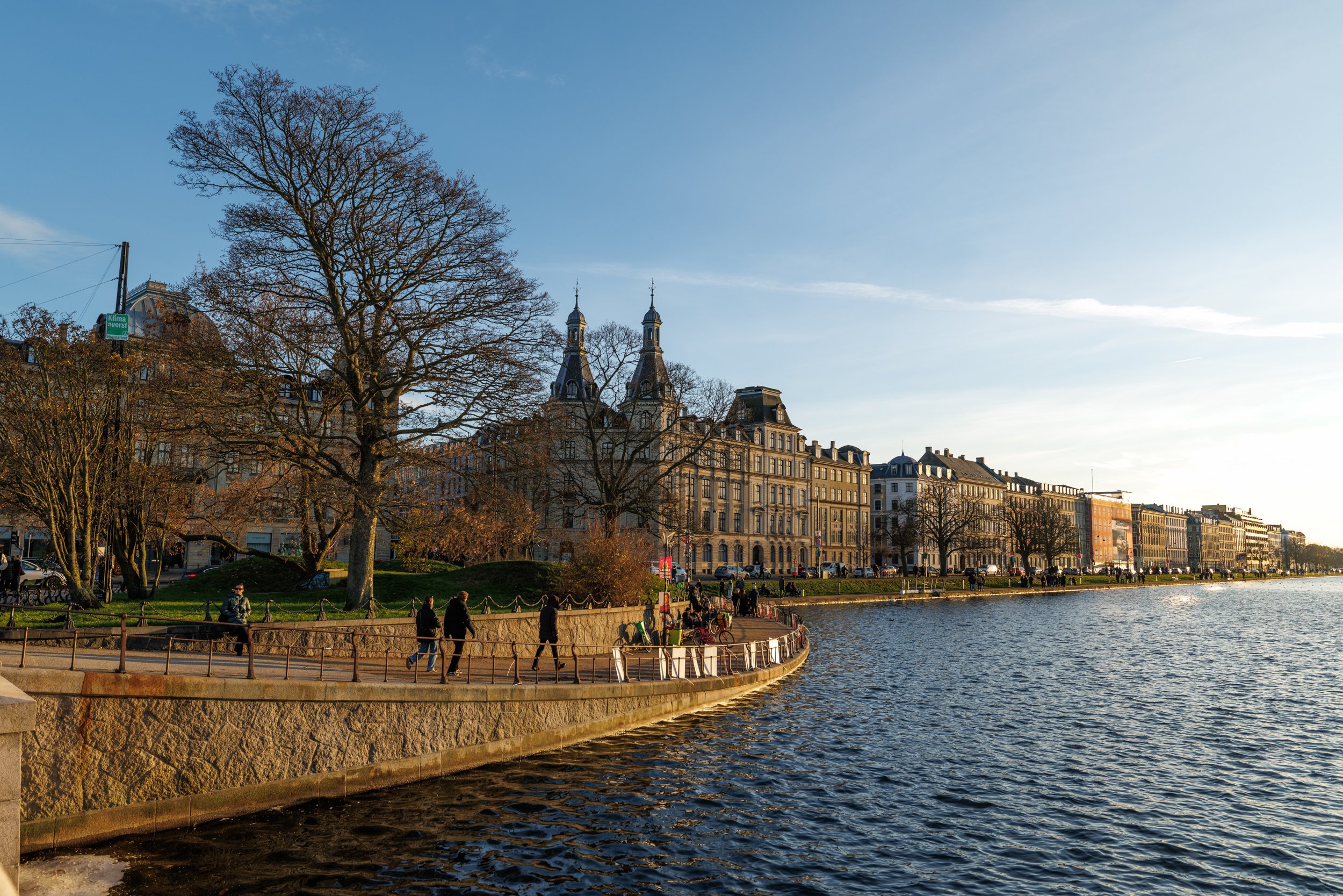 View of a cityscape along a river with historic buildings, leafless trees, and a promenade with people walking and sitting by the water, under a clear blue sky.
