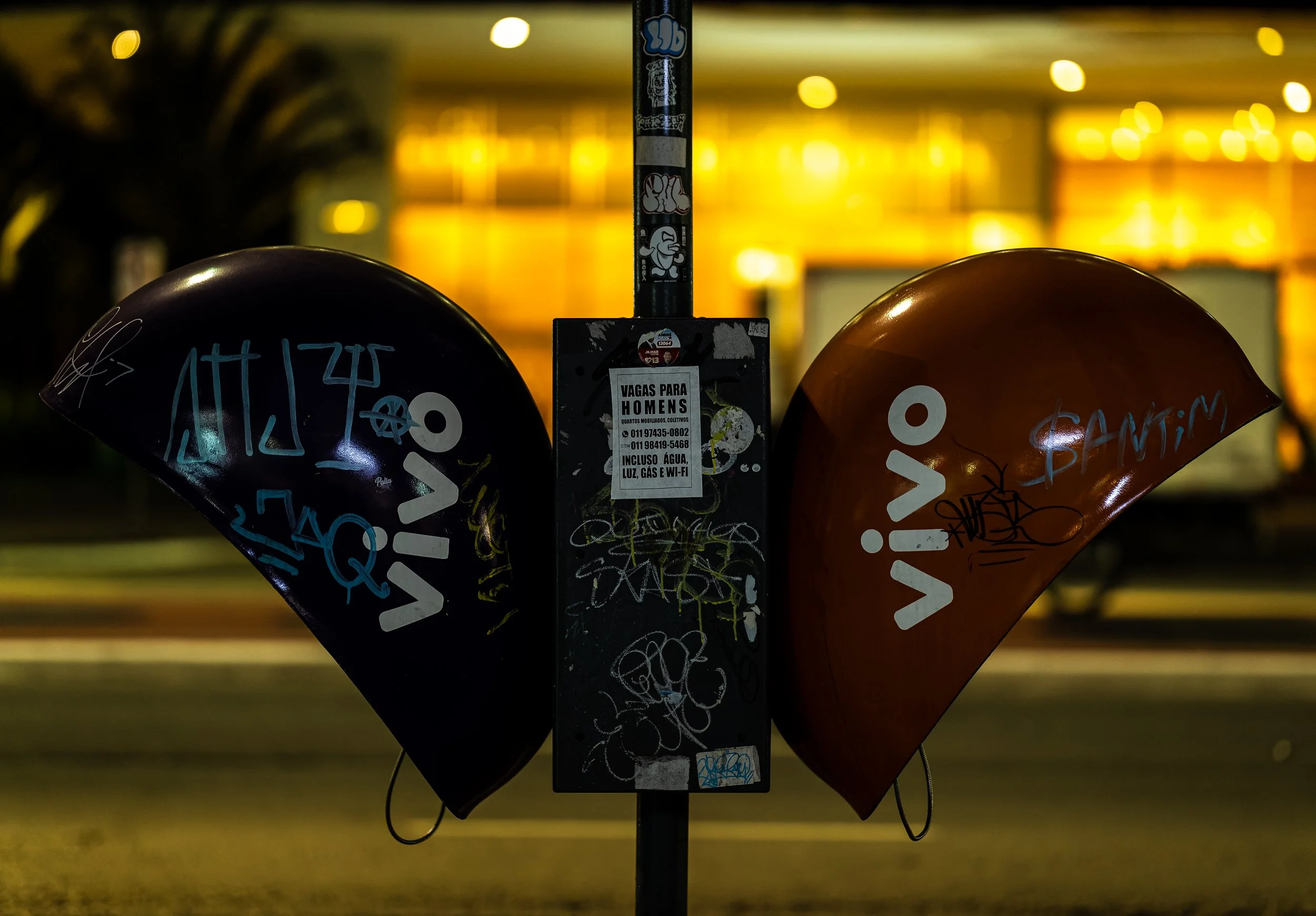 A parking meter with two bike helmets hanging on either side. The helmets are covered in graffiti and markings. The background is blurred with yellow lights, indicating it is nighttime.