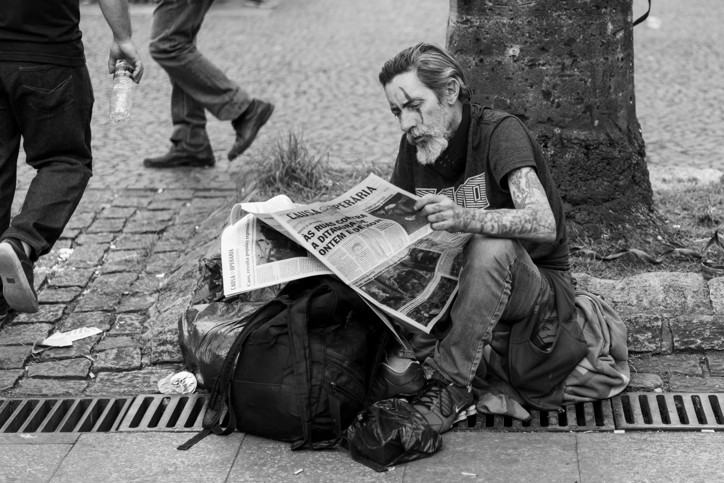 A homeless man with a beard and tattoos, sitting on the sidewalk next to a tree, reading a newspaper with Portuguese language headline.