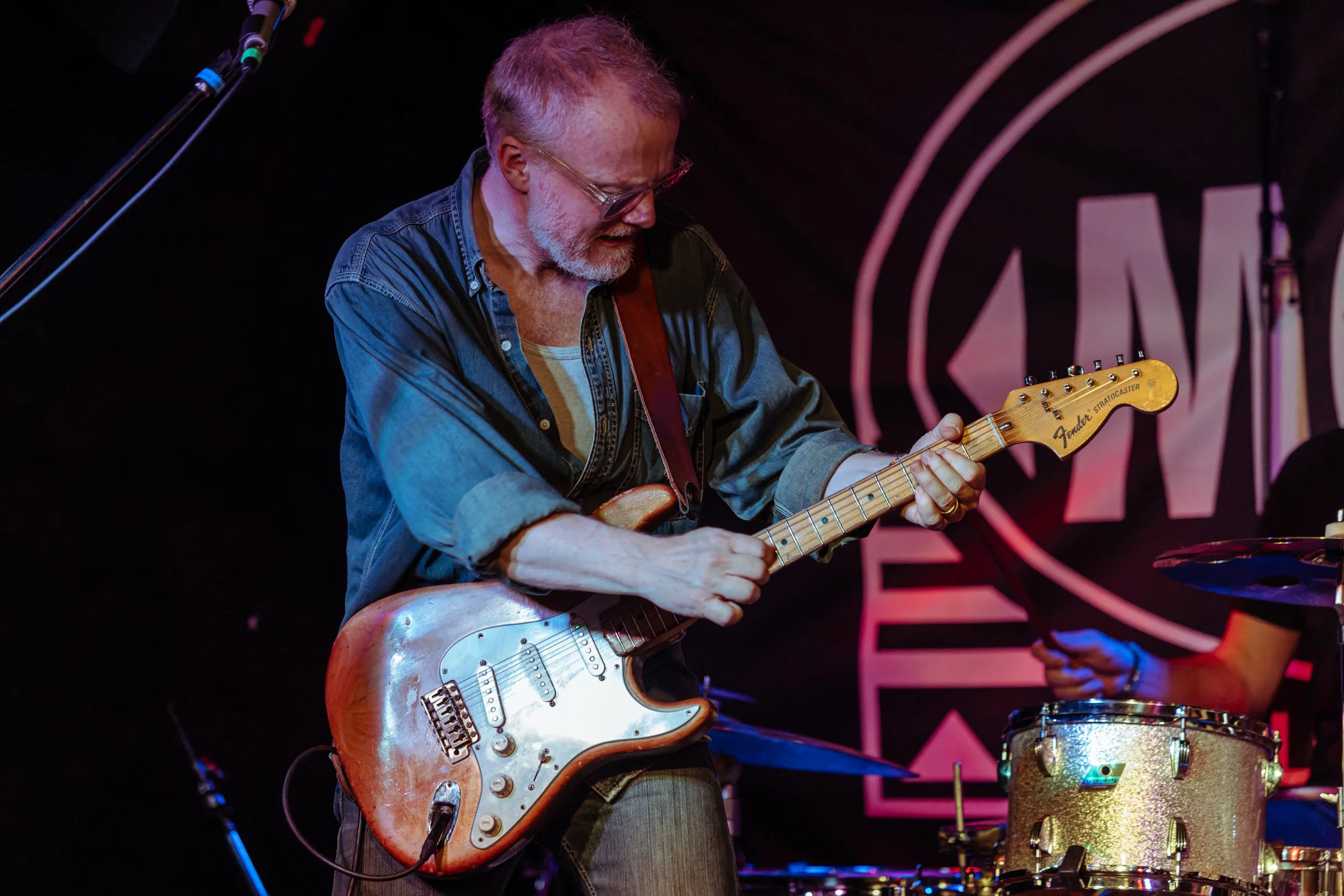 A man with gray hair and glasses playing an electric guitar on stage, wearing a denim shirt and striped t-shirt, with drums visible in the background.
