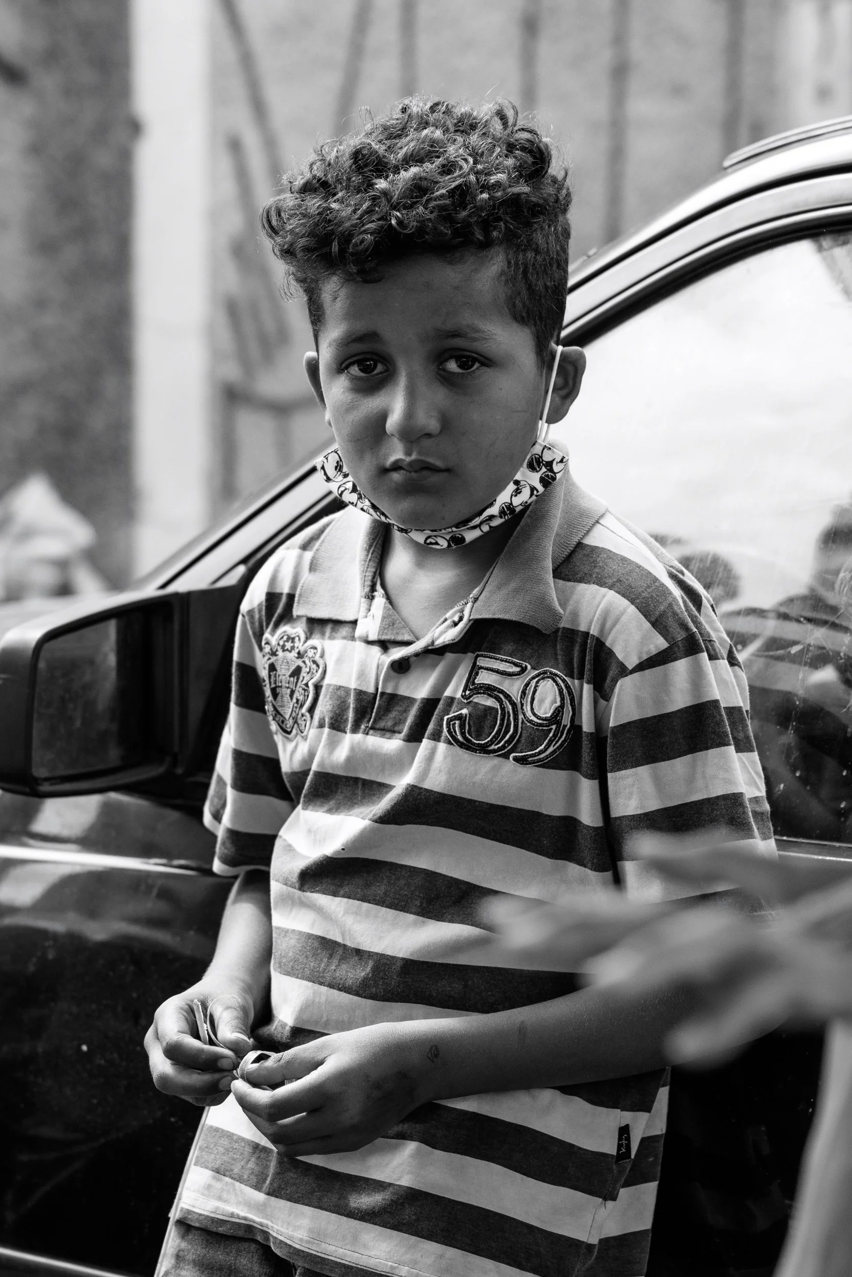 A young boy with curly hair and a serious expression wearing a striped polo shirt, standing outdoors near a parked car.