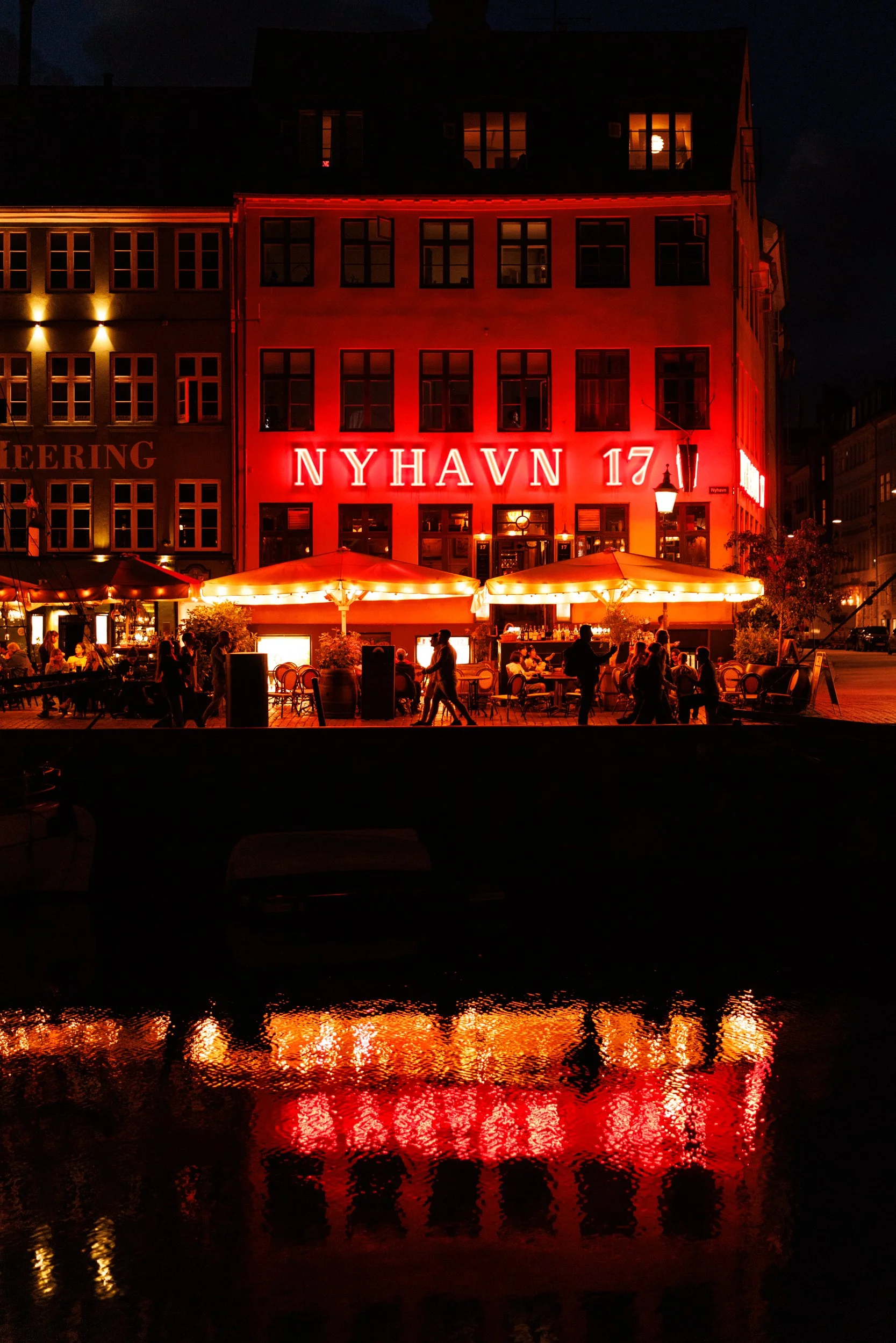 Nighttime cityscape with a building lit up in red neon lights reading 'NYHAVN 17,' a waterfront with reflections of the lights, outdoor seating with umbrellas, and people walking.