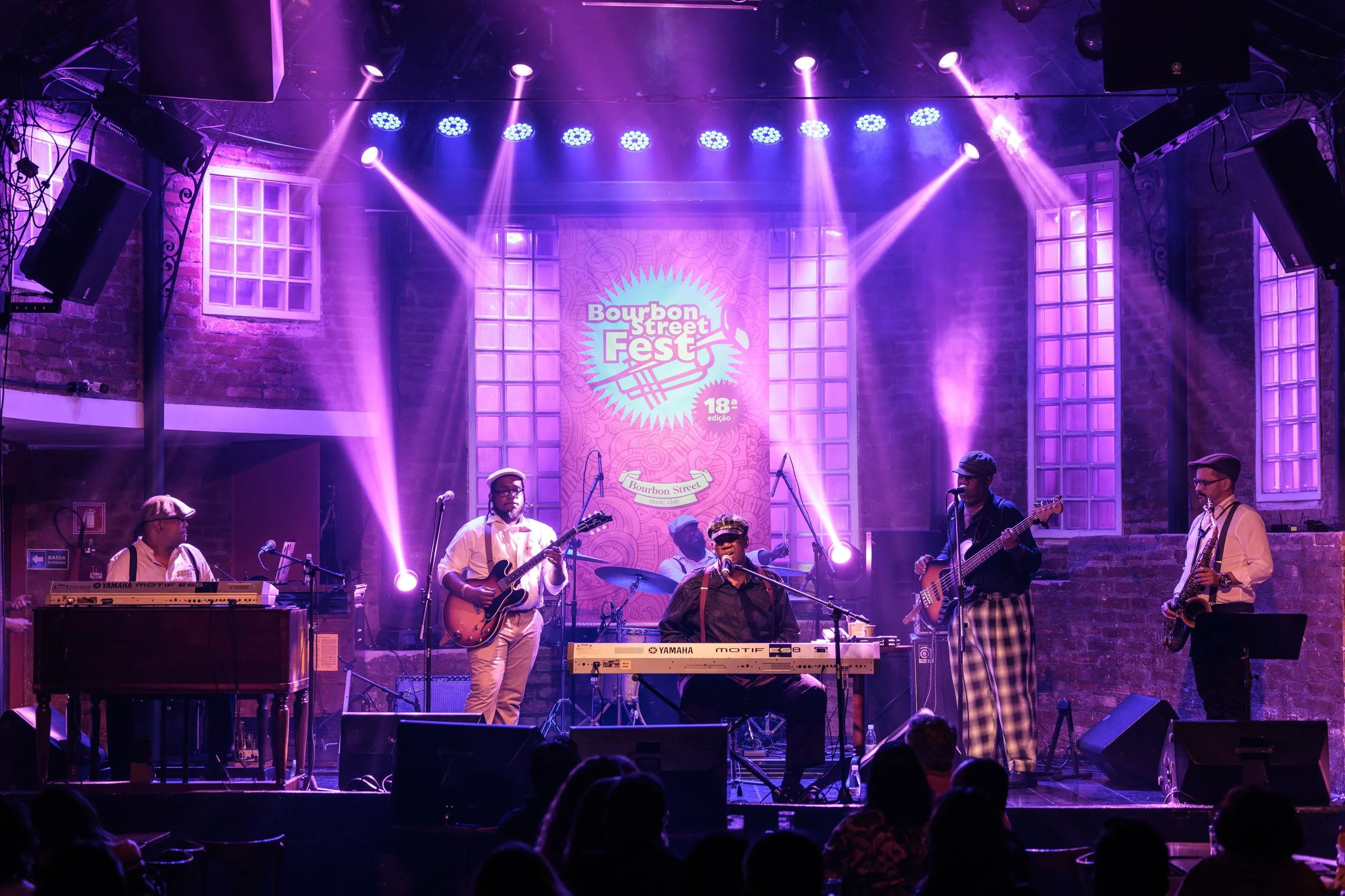 Musicians performing on stage at Bourbon Street Fest, with purple lighting and a large banner behind them. The stage has multiple instruments and microphones, and is set inside a venue with brick walls and large windows.