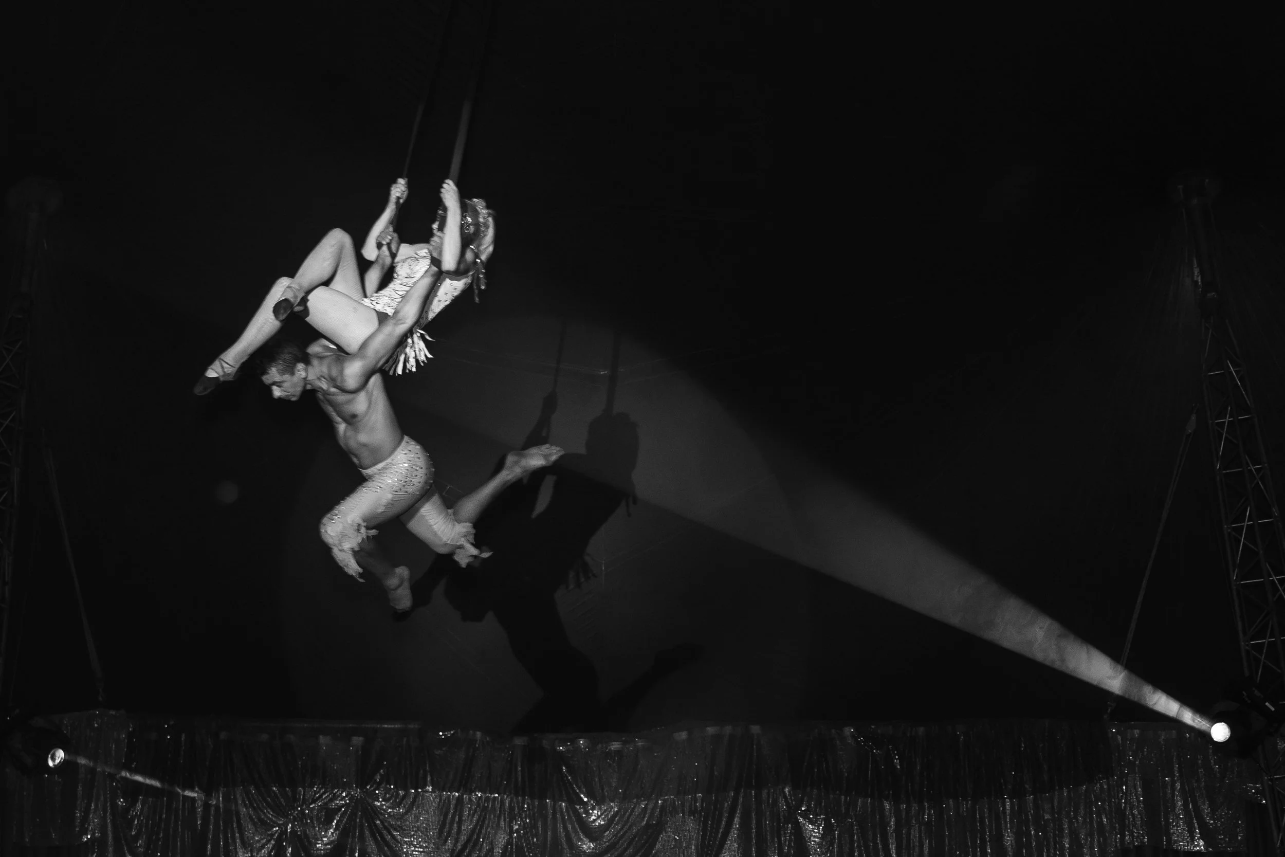 A performer is hanging upside down on a trapeze, holding a woman wearing a dress with a floral pattern, in a circus or performance setting.