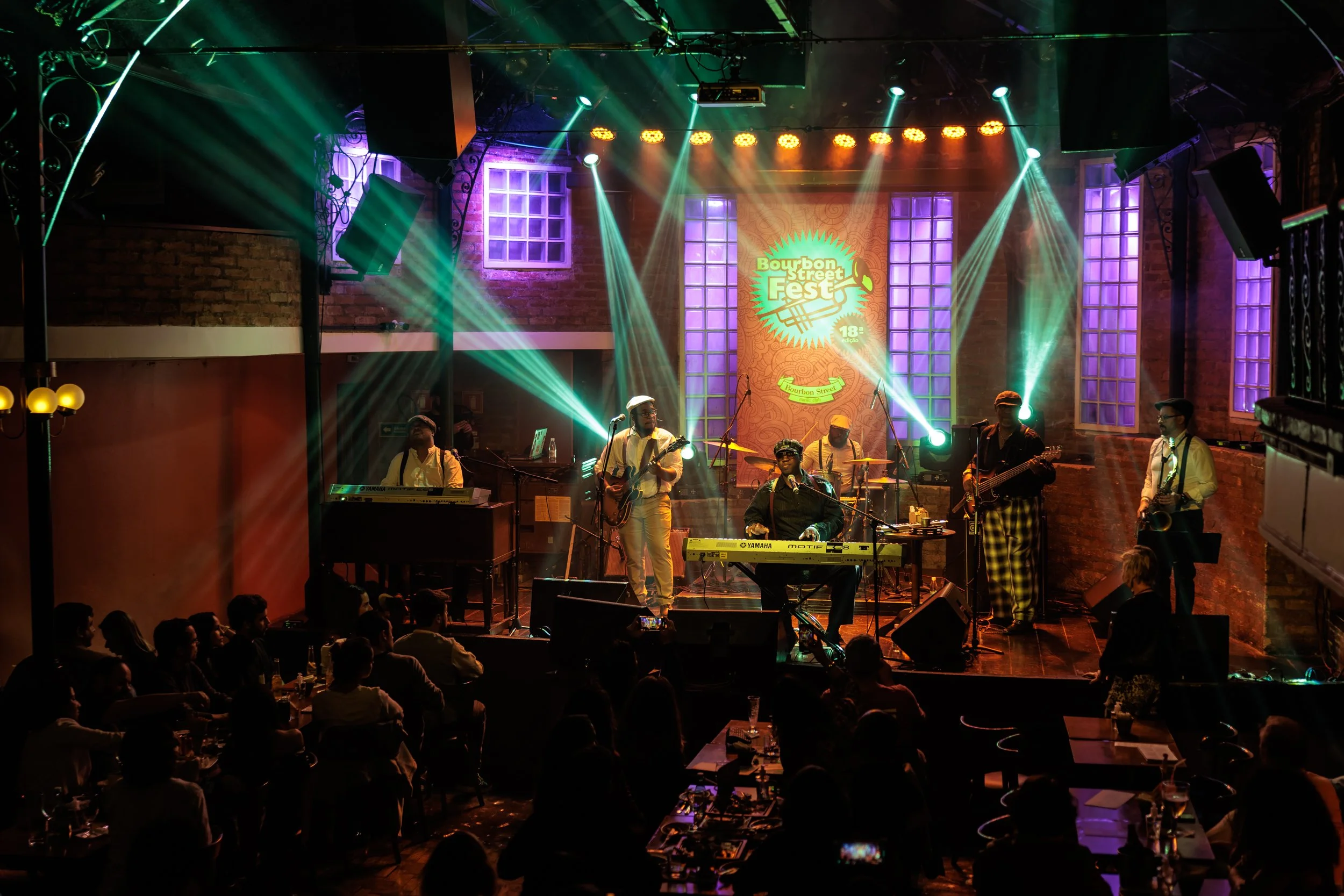 Live music performance at Bourbon Street Fest, with a band playing on stage under colorful stage lights, audience seated in front, and a backdrop with the event logo.