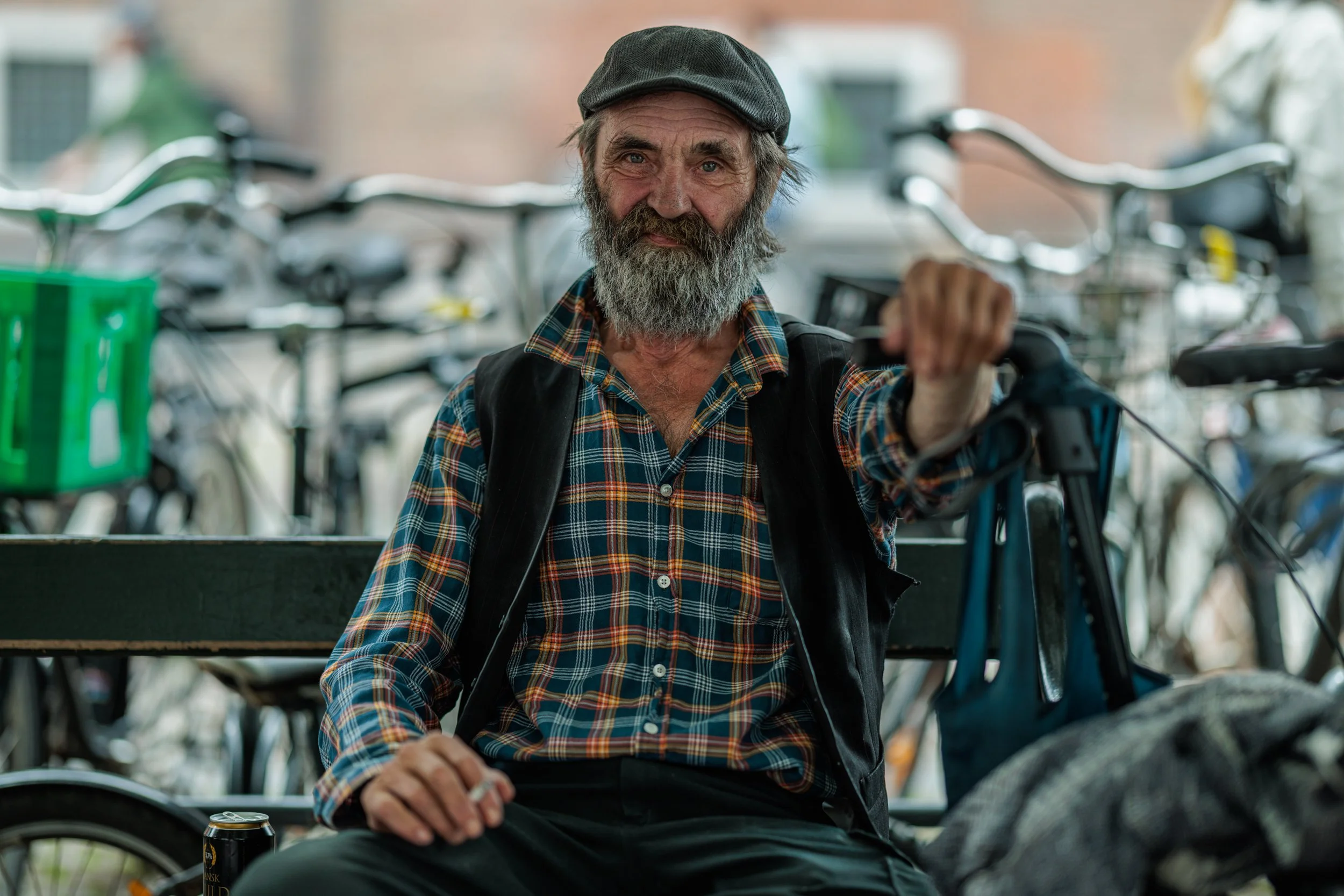 An elderly man with a gray beard and mustache, wearing a dark flat cap, plaid shirt, and black vest, sitting on a bench in front of a bicycle shop with multiple bicycles in the background. He has a slight smile and is holding a soda can in his right 
