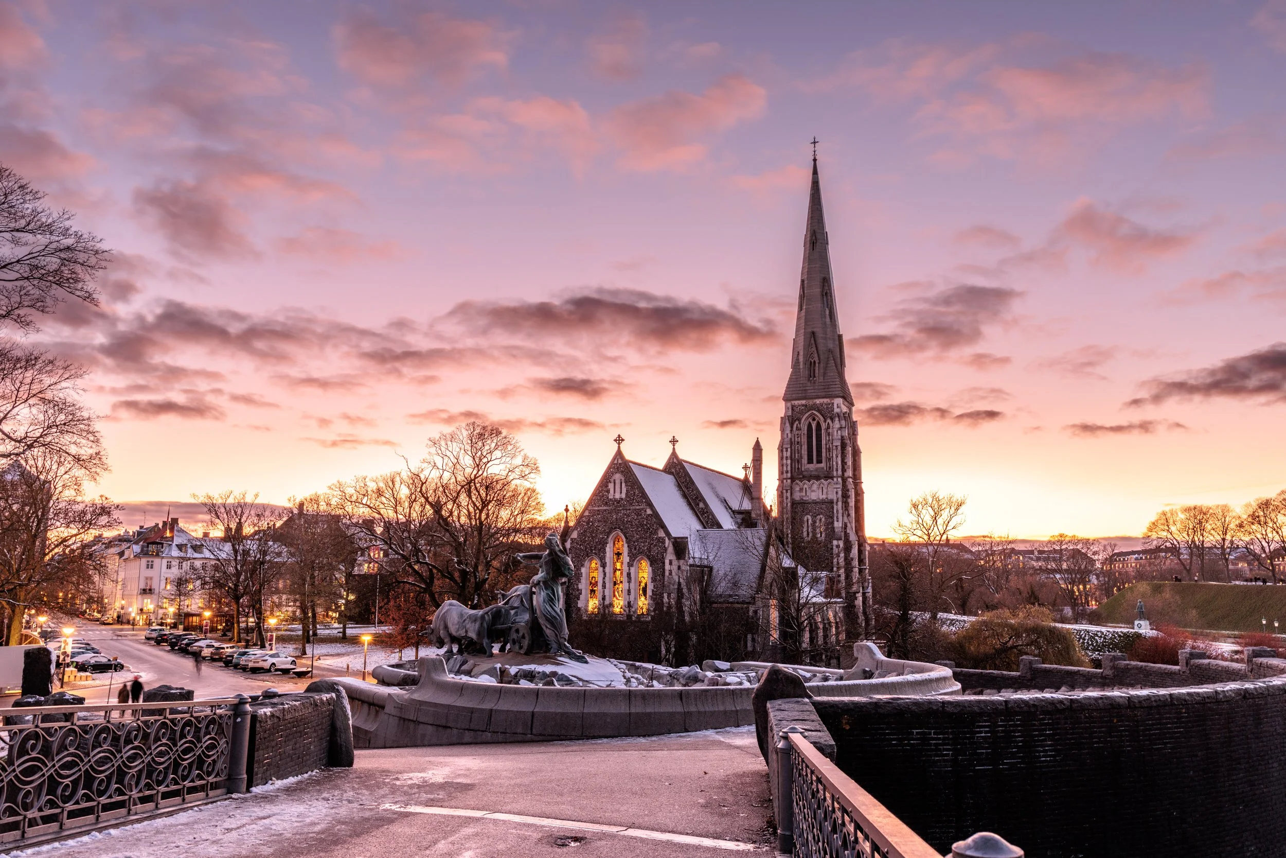 A church with a tall steeple towards the center, surrounded by leafless trees and a cloudy evening sky with pink and purple hues. A fountain with sculptures is in the foreground, and city buildings with lights are visible in the background.