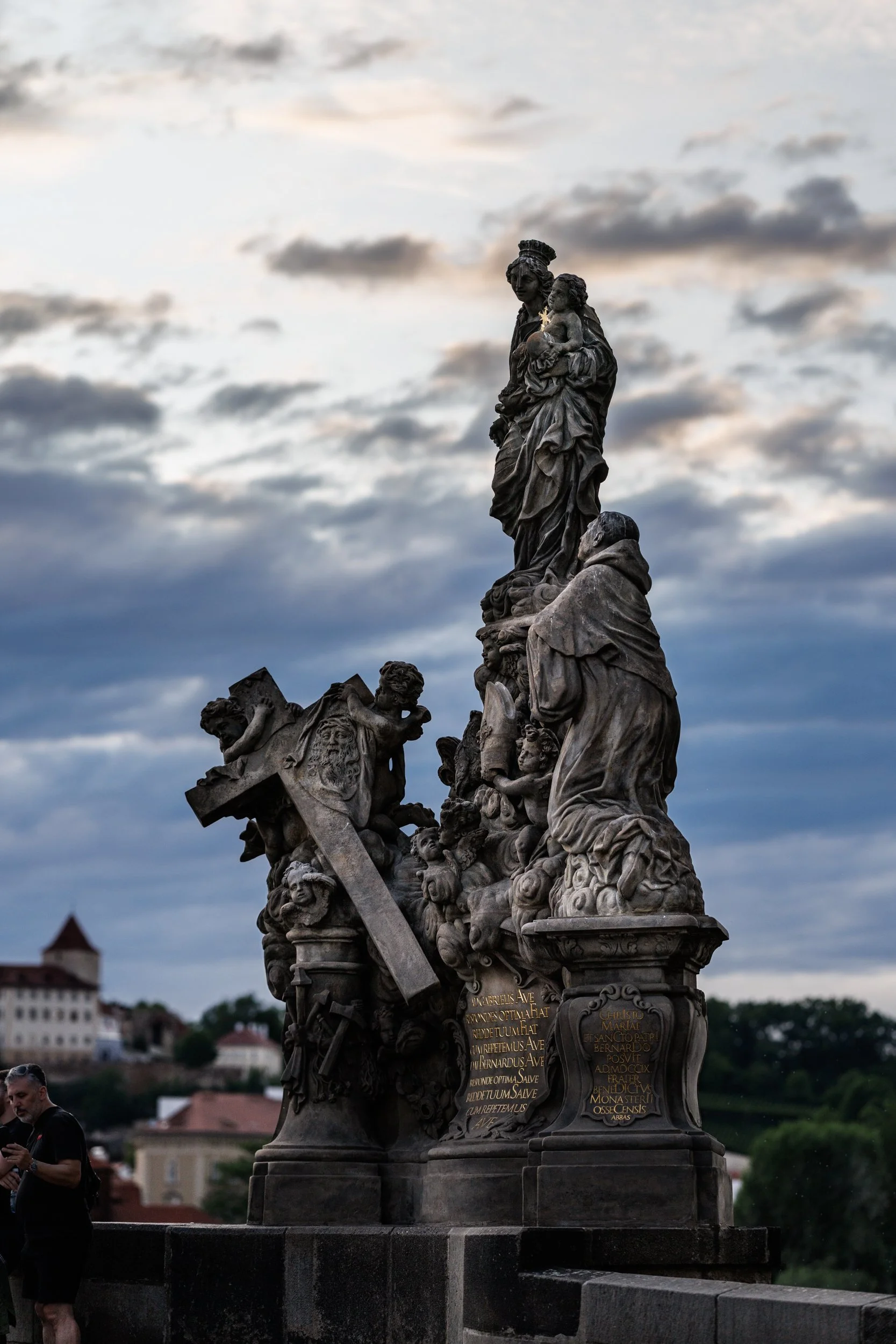 A detailed stone sculpture of religious figures and decorative elements, with engraved Latin inscriptions, set against a cloudy sky and distant buildings.
