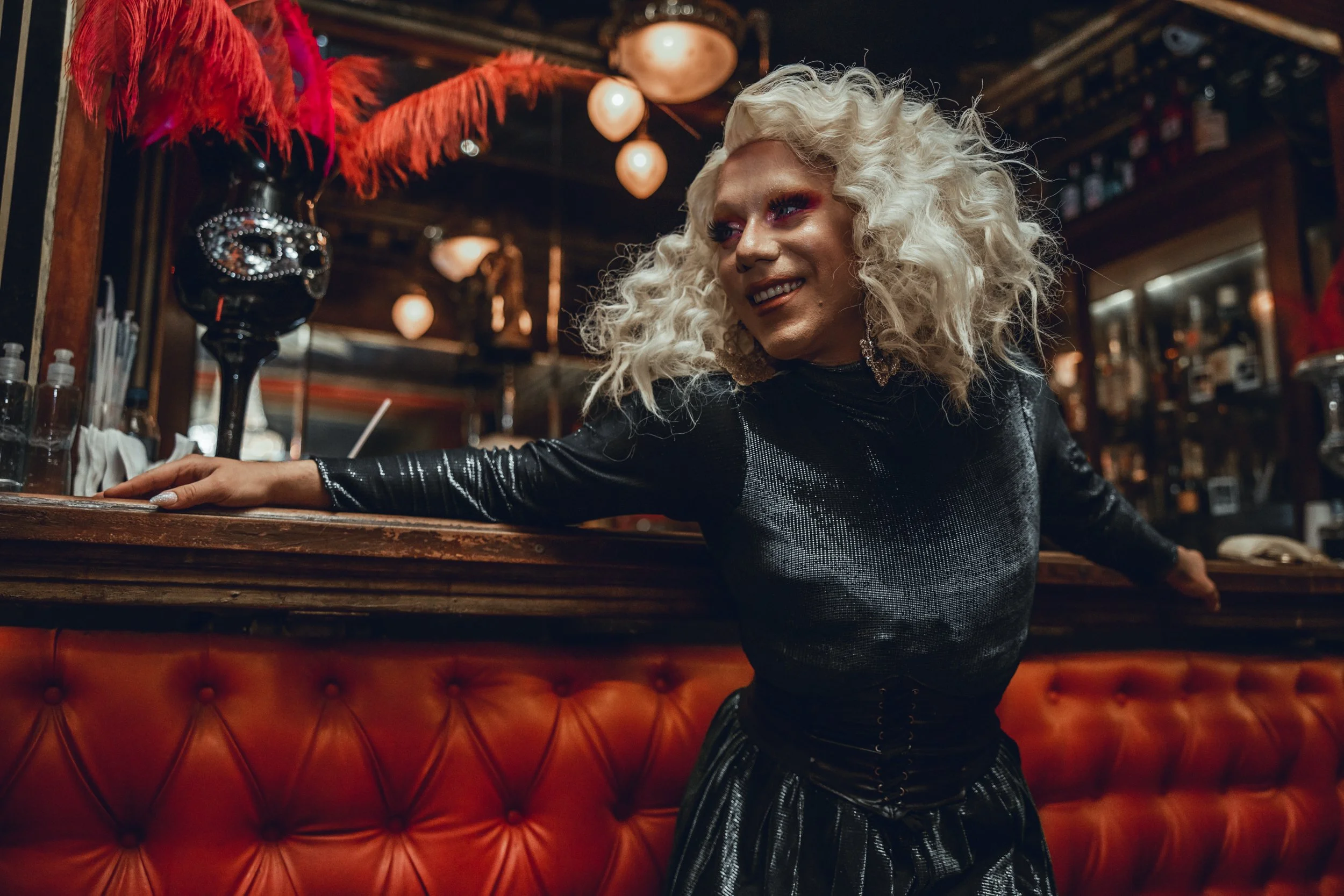 A person with curly platinum blonde hair and pink eye makeup leaning on a bar counter in a dimly lit bar. They are wearing a shiny black long-sleeve top.