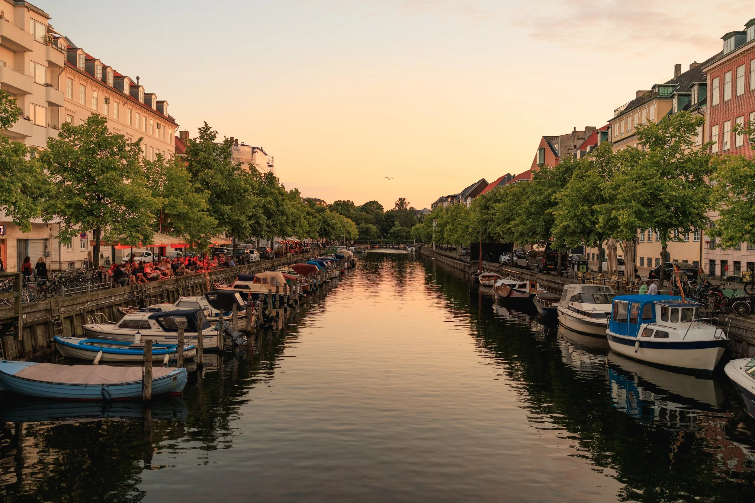 A canal scene at sunset with boats docked along the sides, outdoor cafes with red umbrellas, and multi-story buildings with trees lining the street.