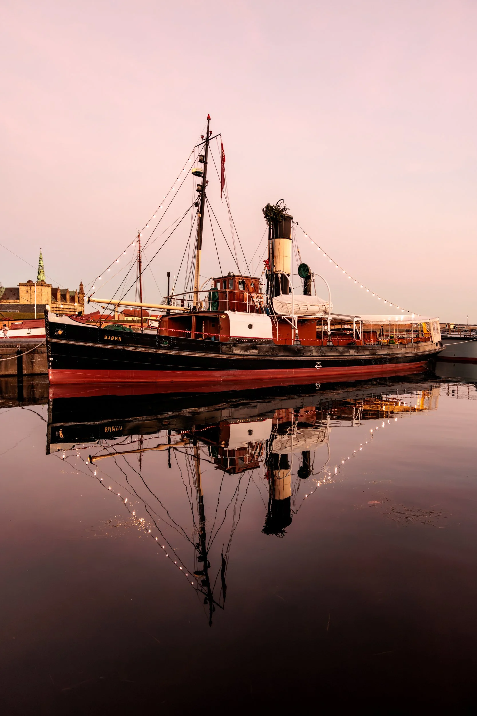 A vintage boat named BJORN docked at a pier during sunset, with calm water reflecting the boat and sky, adorned with string lights and a cityscape with historic buildings in the background.