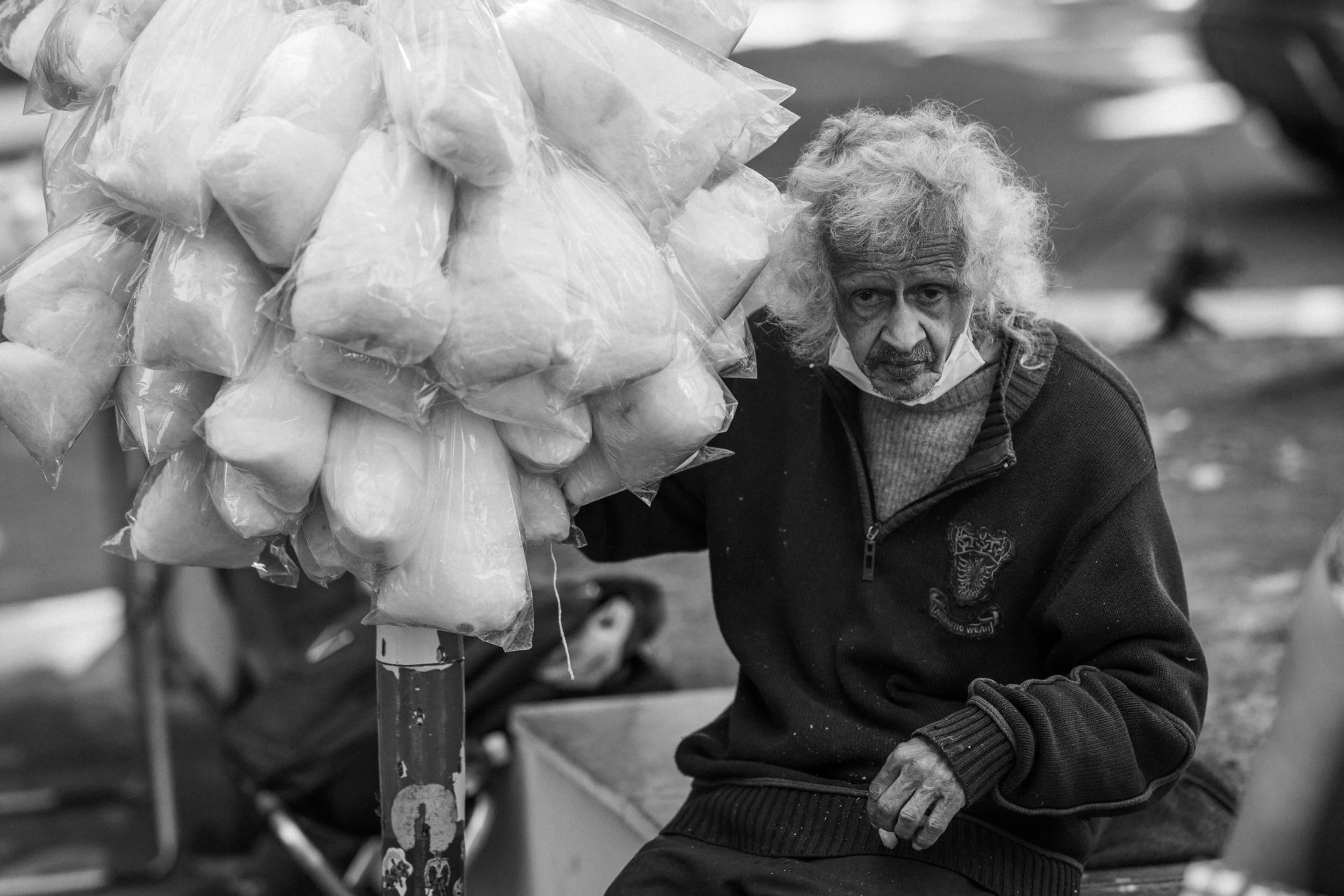 An elderly man with white curly hair, wearing a dark zip-up jacket, is holding a large bag of cotton balls, which is resting on a stand. The man is looking directly at the camera with a serious expression.