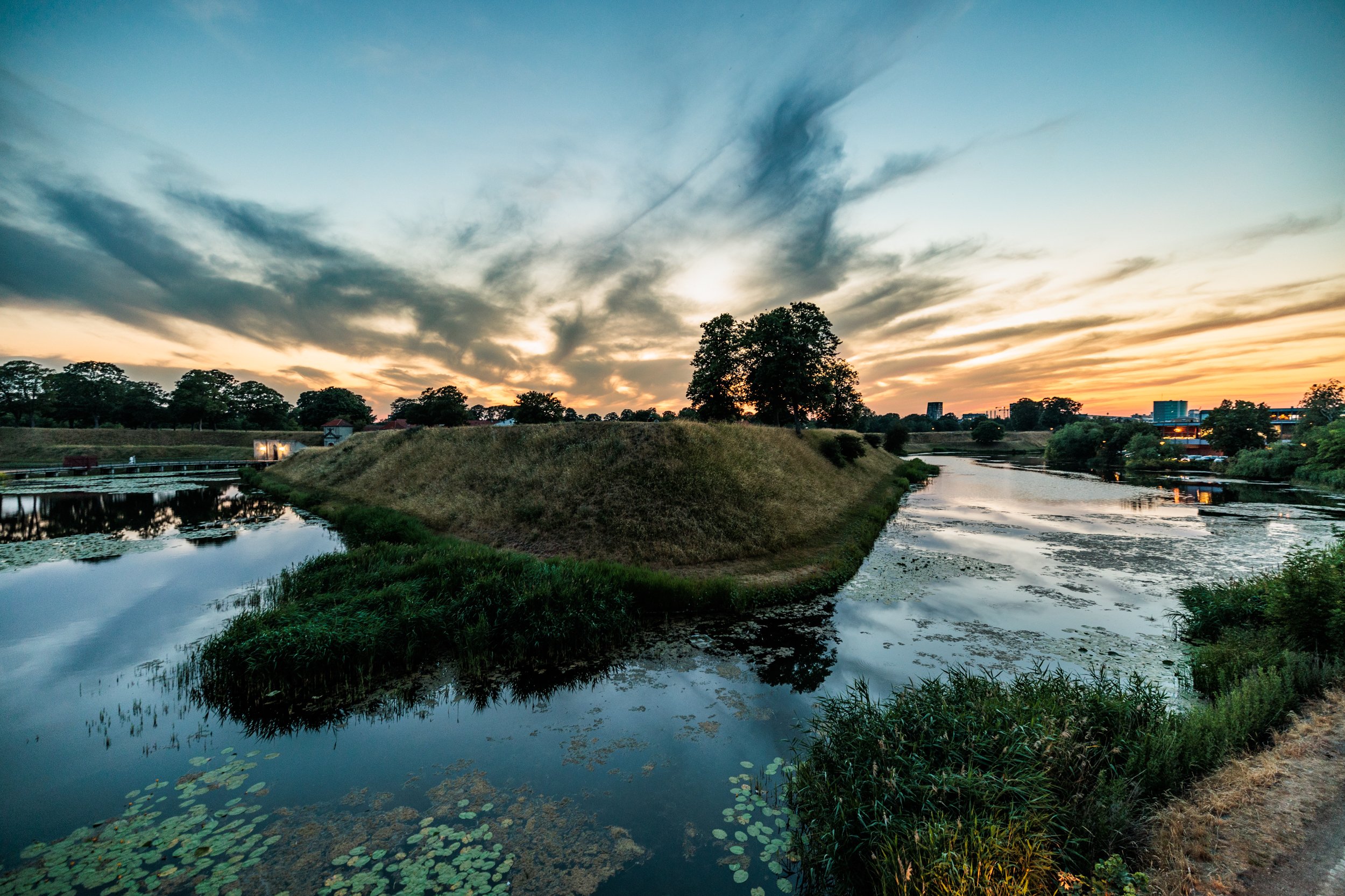 A river flowing through a natural landscape at sunset with trees, grassy banks, and a cloudy sky reflected in the water.