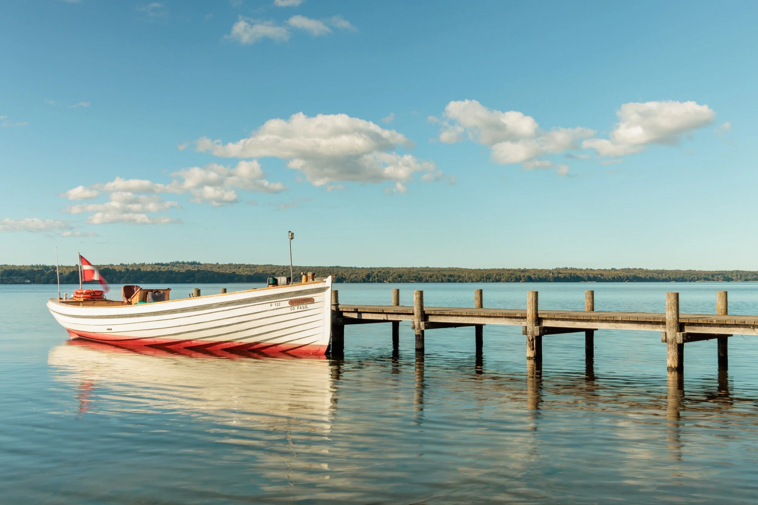 A small boat tied to a wooden dock on calm water, with a Danish flag at the stern and a partly cloudy sky overhead.