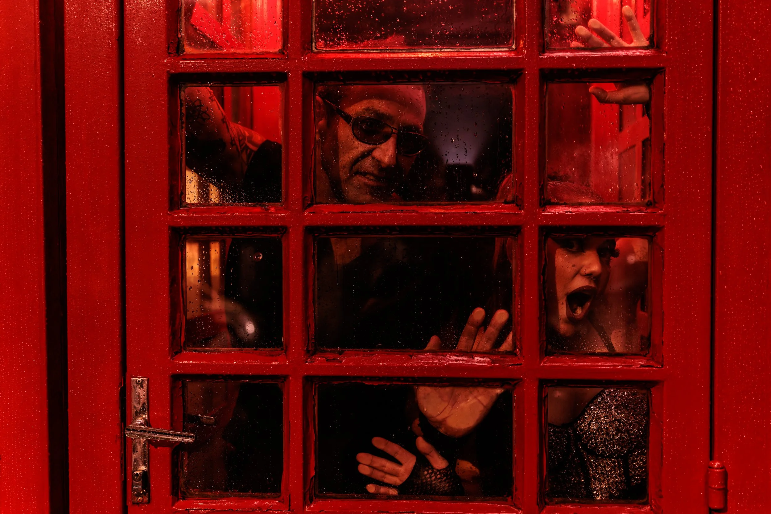 People behind a red telephone booth, one person wearing sunglasses, another with an open mouth and hand raised, rain on the glass.