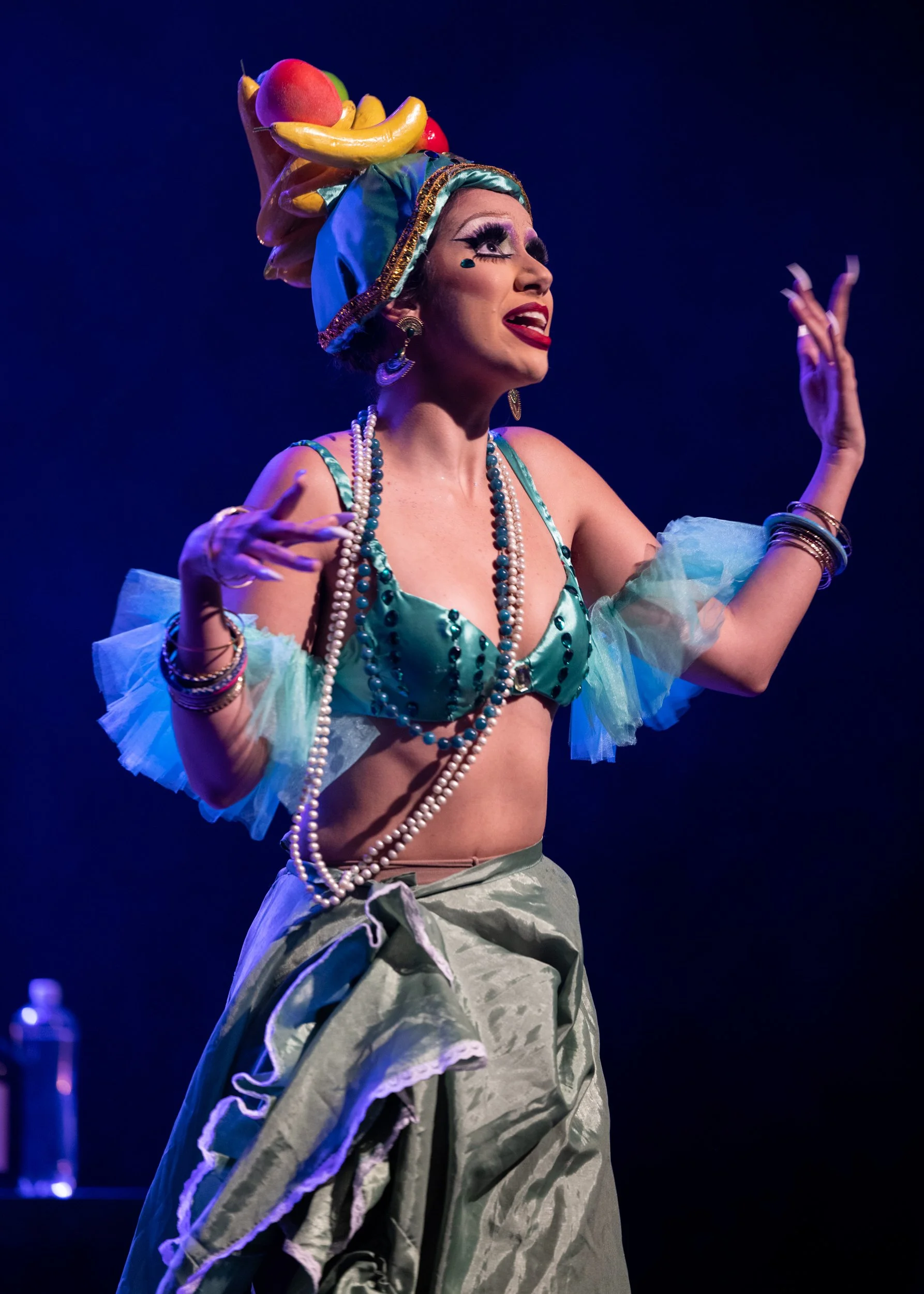 Performer on stage wearing a colorful fruit-themed headdress, extravagant makeup, jewelry, and a semi-transparent costume against a dark background.
