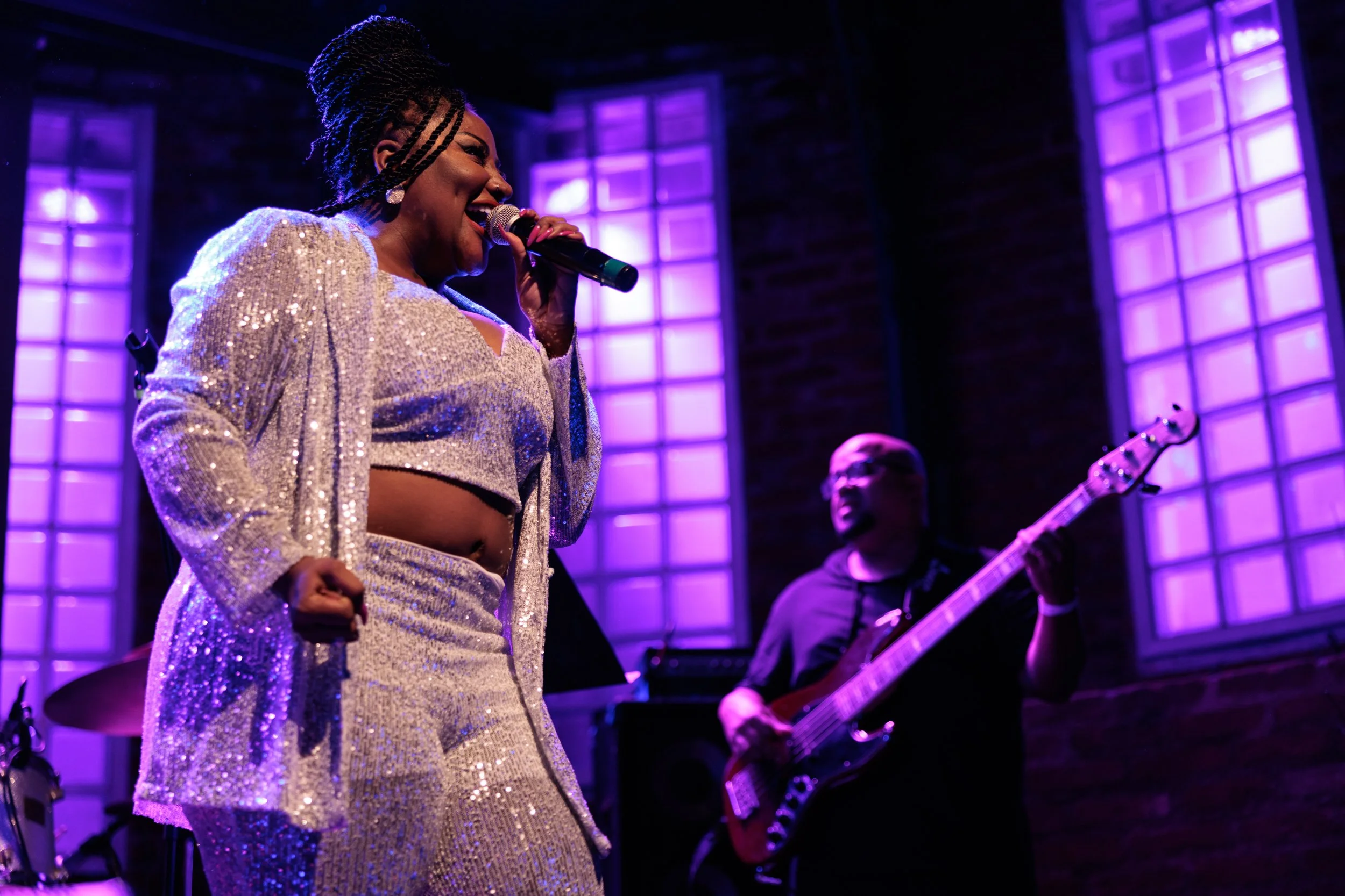 A female singer with braided hair, wearing a shimmering silver outfit, performs with a microphone on stage. Behind her, a guitarist wearing glasses plays an electric guitar. The background features purple-lit windows and brick walls, creating a concert or live music venue atmosphere.