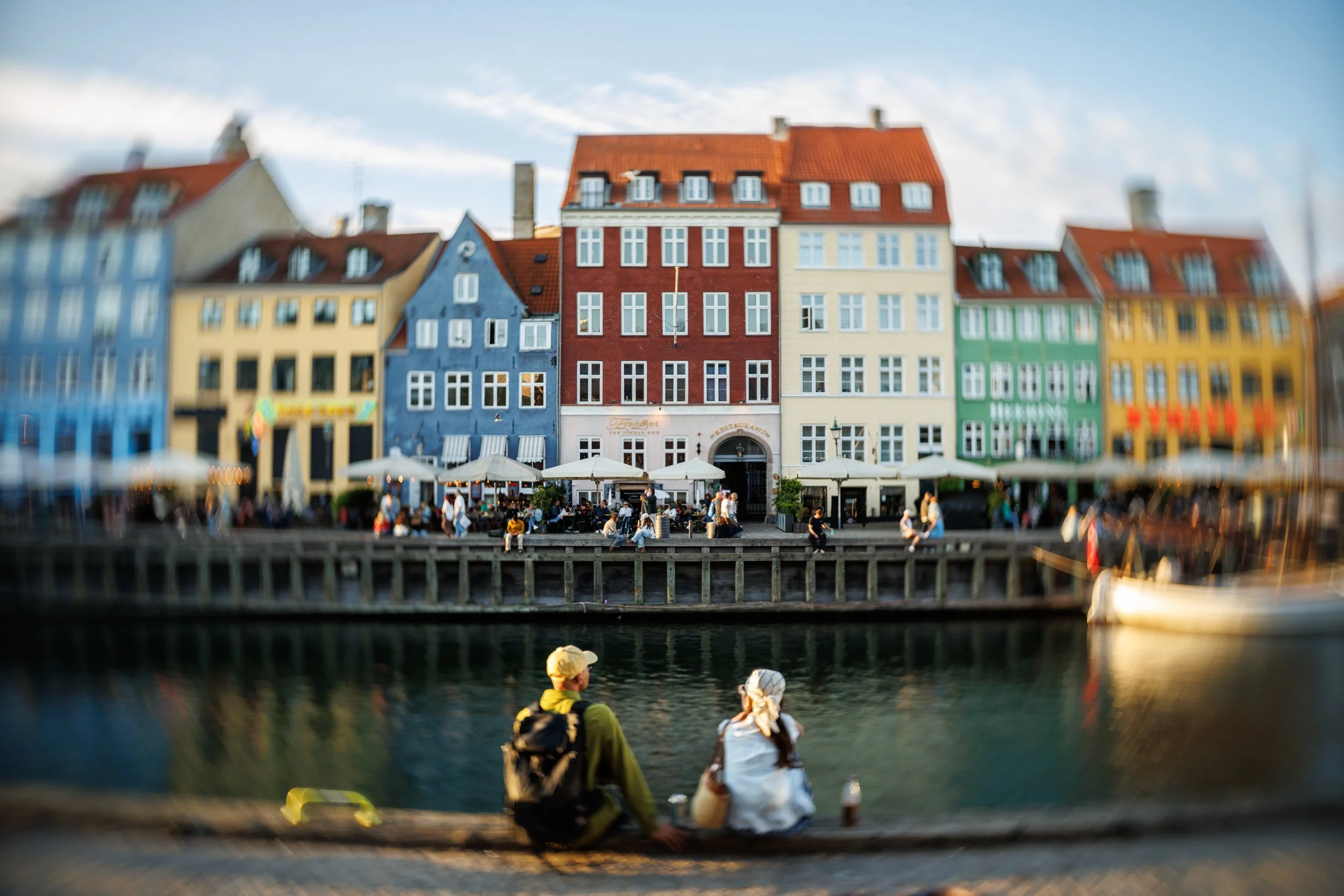 Colorful European buildings with outdoor cafes along a waterfront, with people sitting by the water and boats docked nearby.