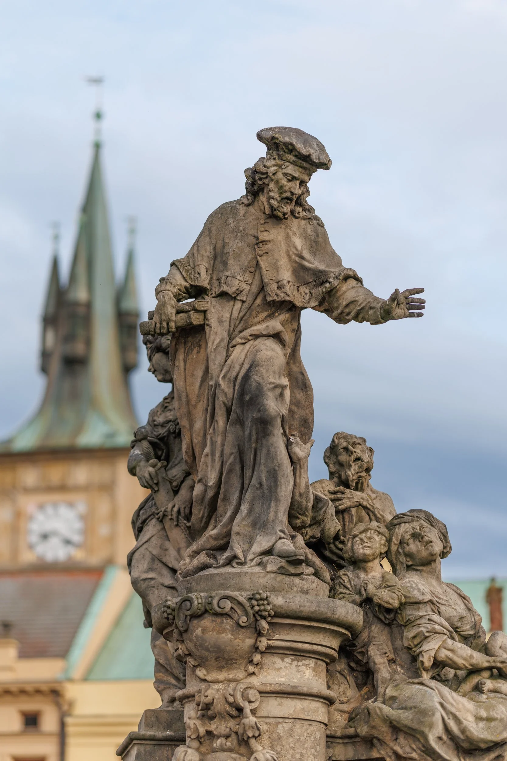 A detailed stone sculpture of multiple figures, including a central male figure with a beard and hat, surrounded by children and other figures, set against a backdrop of a historic building with a clock tower and spires.