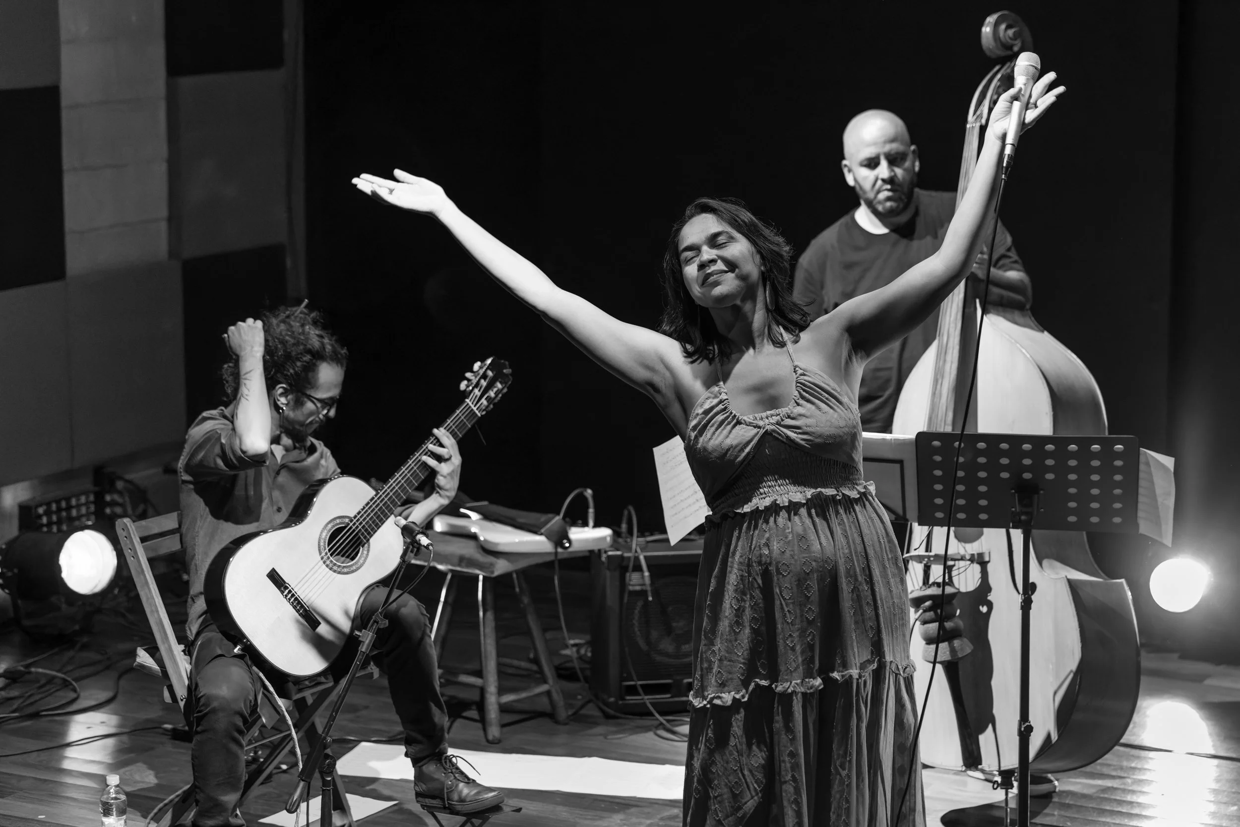 A woman singing with arms outstretched, a guitarist playing, and a man with a double bass on stage during a performance.