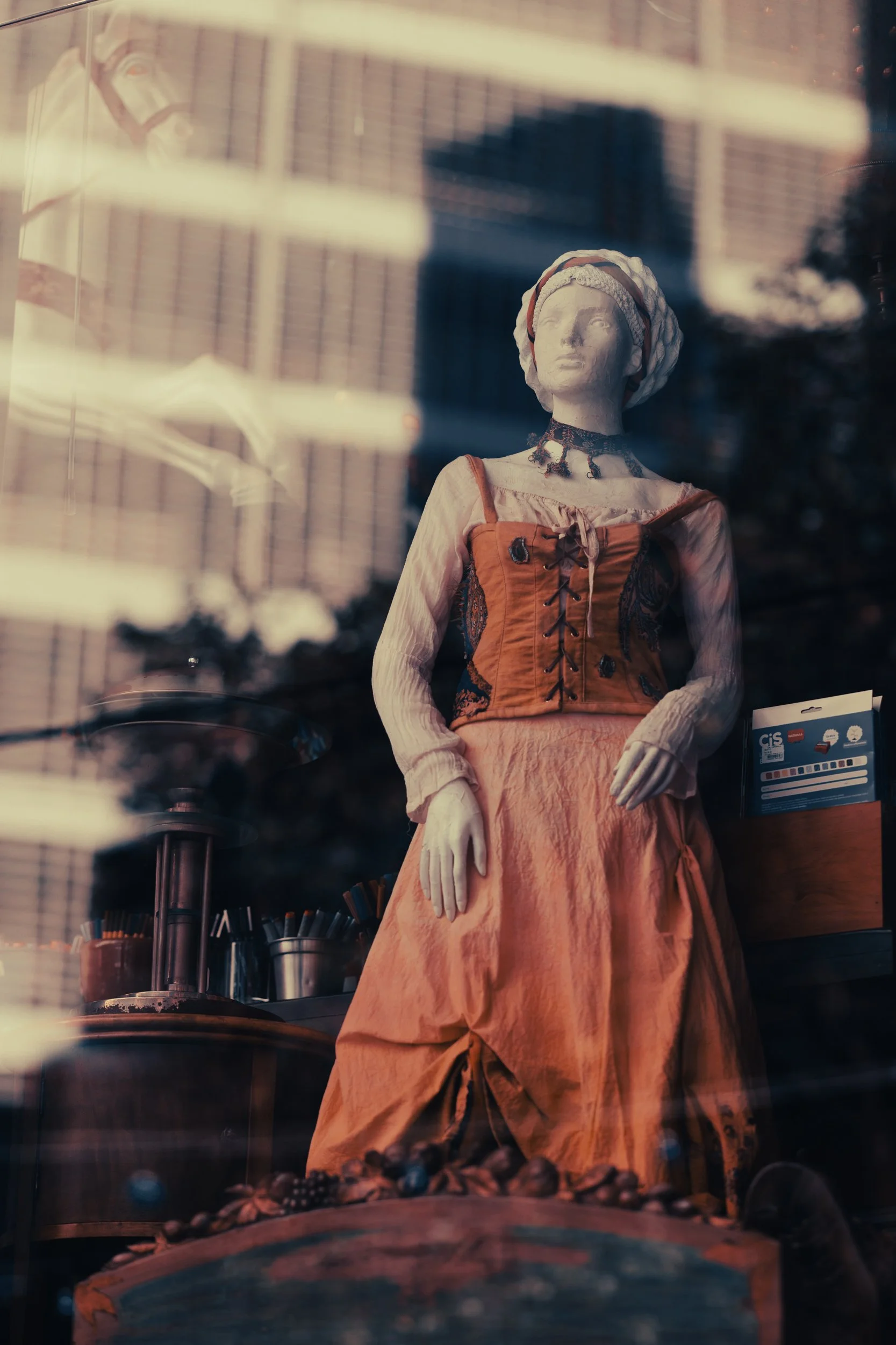 A mannequin dressed in a vintage style outfit, featuring an orange corset, long sleeves, and a pink skirt, placed in a store window with reflective glass, showing blurred reflections and surrounding store items.