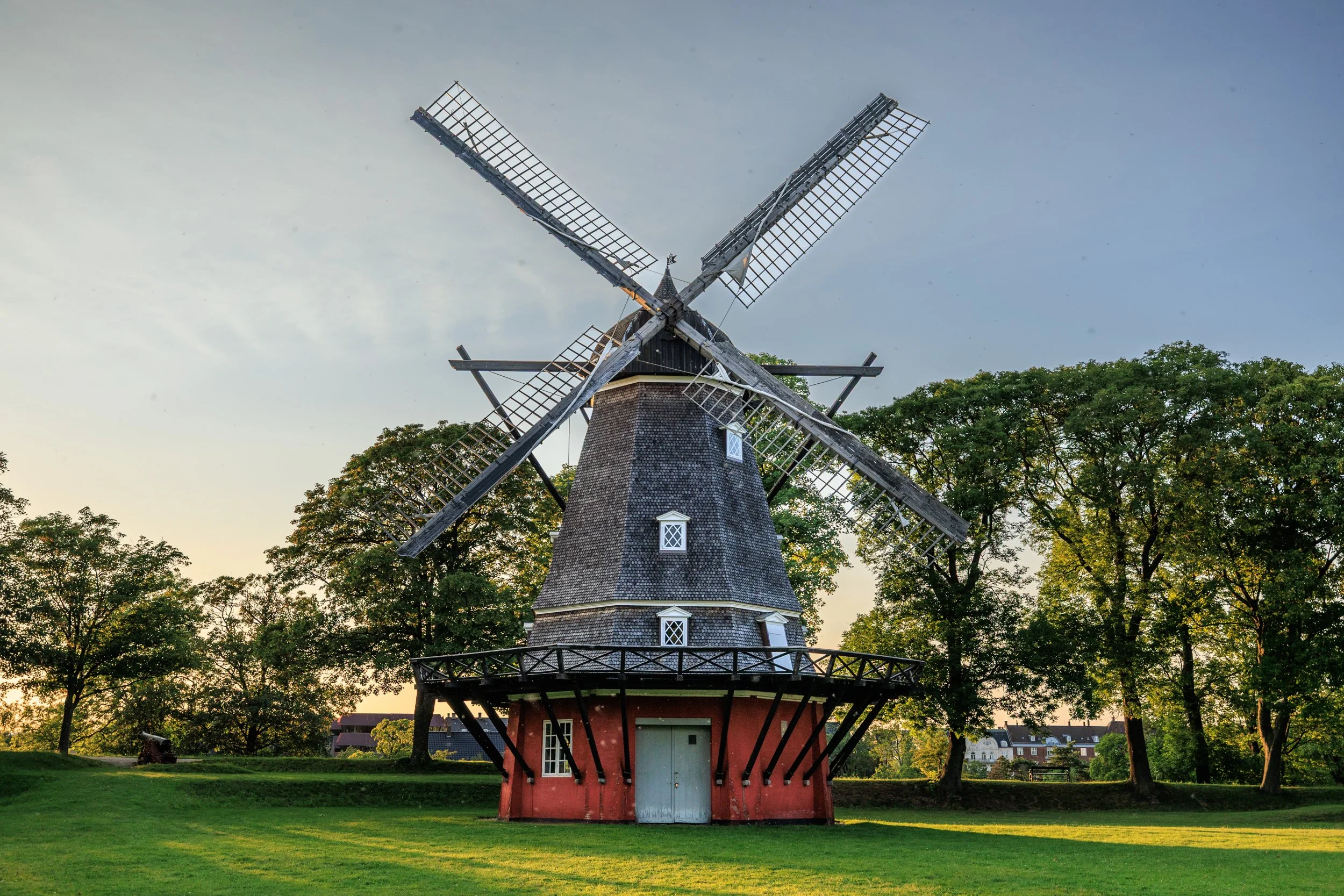 A traditional windmill with wooden blades and a red base, surrounded by green trees and grass, under a clear sky.