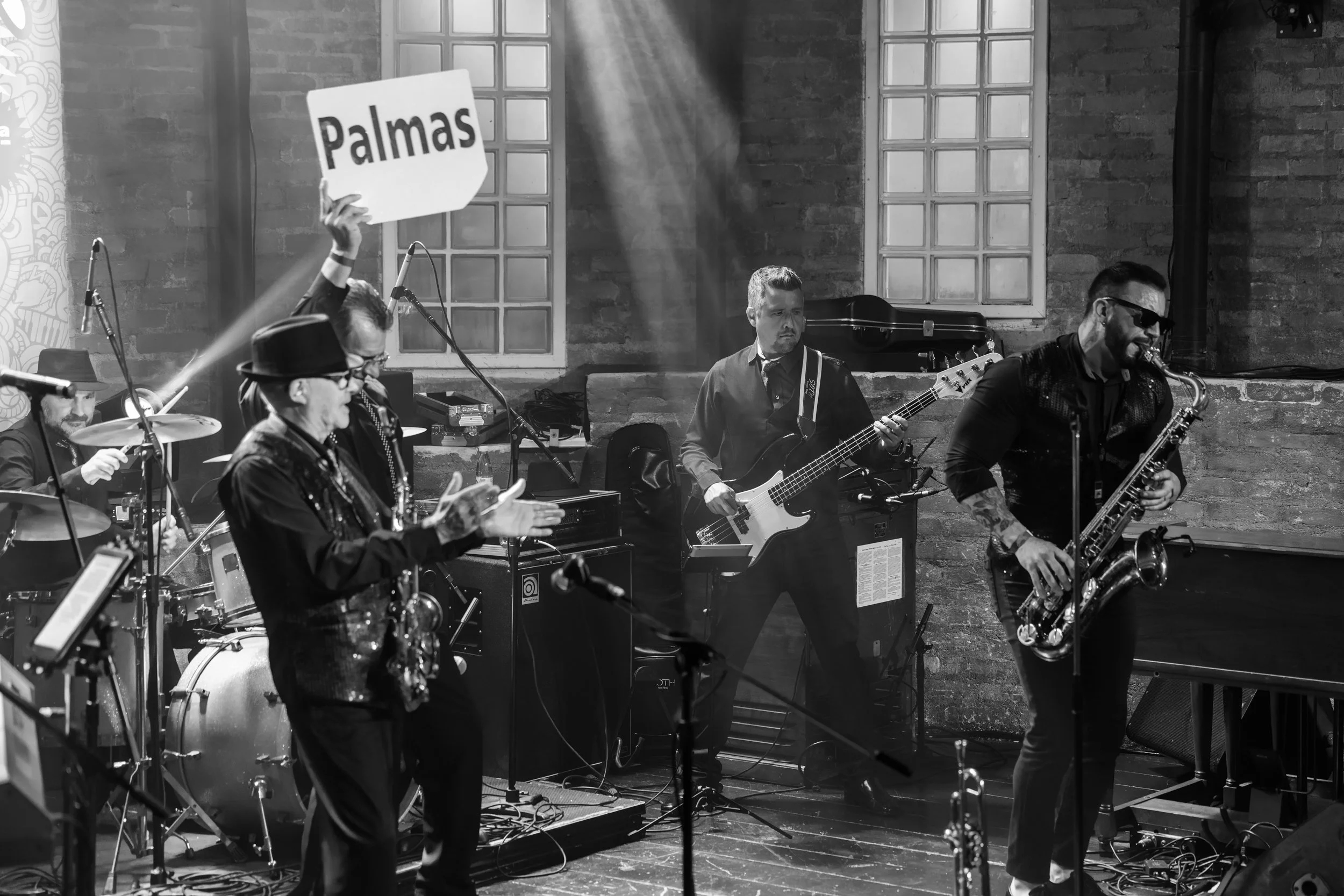 A band performing live on stage at an indoor venue with brick walls and large window panels, featuring musicians playing drums, bass guitar, and saxophone, with one person holding a sign that says 'Palmas'.
