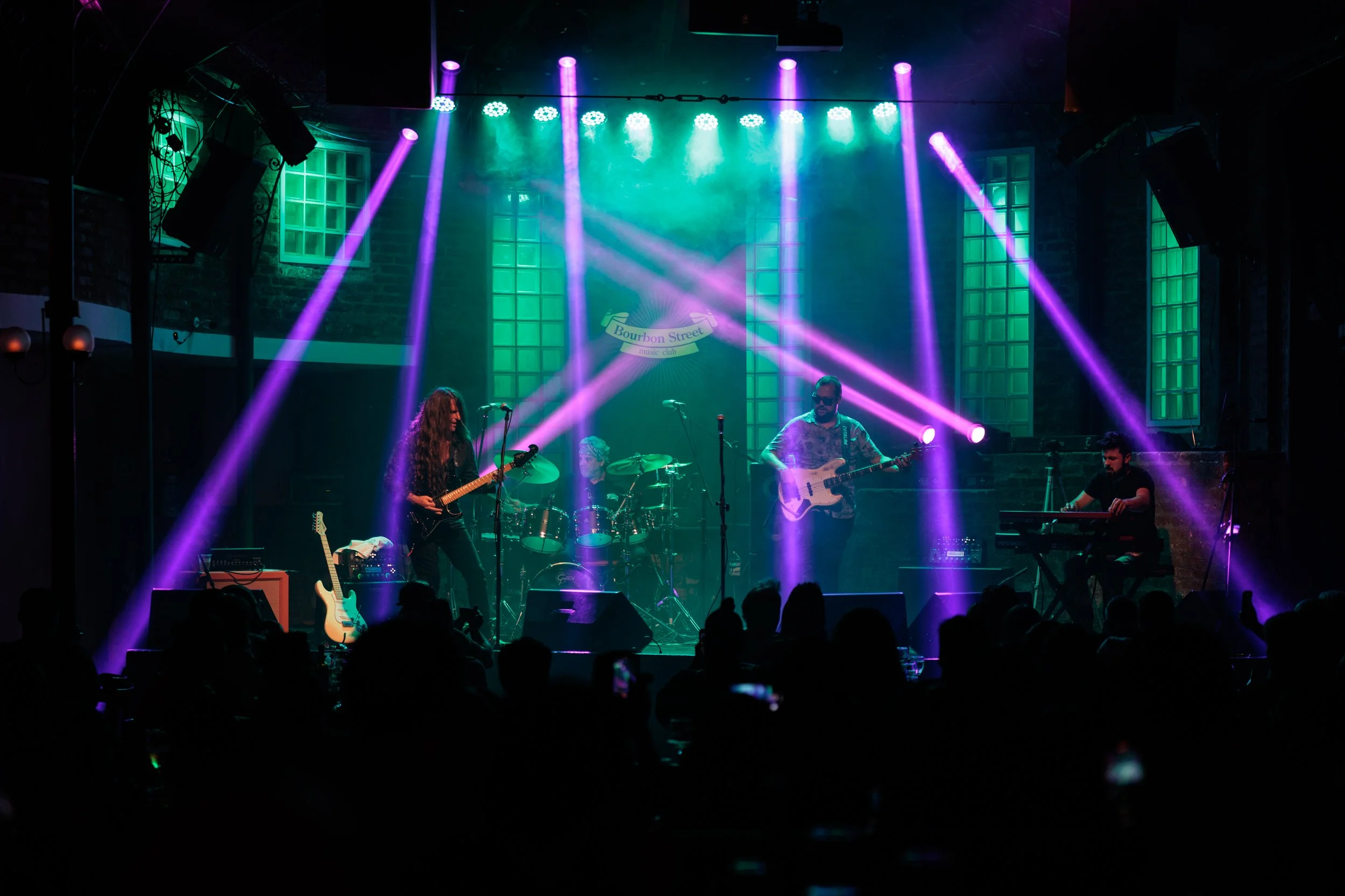 A band performs on stage at Bourbon Street music club, with colorful stage lights and music equipment, audience visible in foreground.