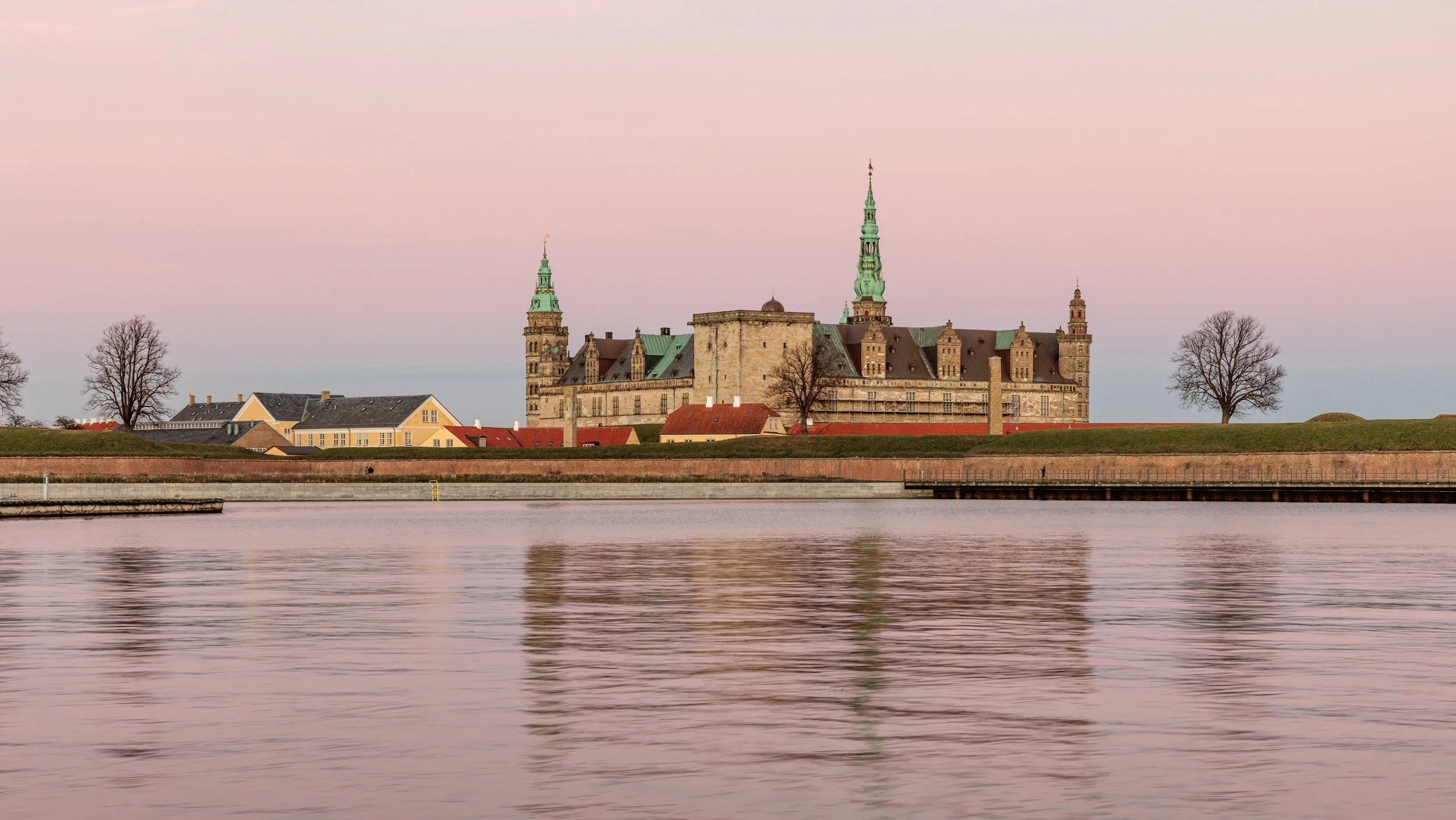 A historic castle with green spires and towers, situated by a body of water with a pinkish sky in the background.